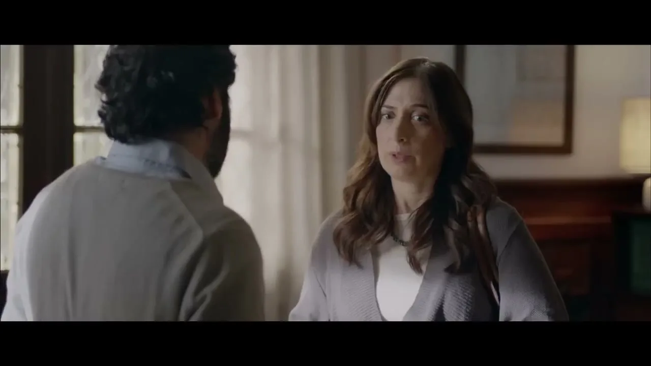A woman with long brown hair and a grey cardigan talks to a man with his back to the camera in a softly lit room.