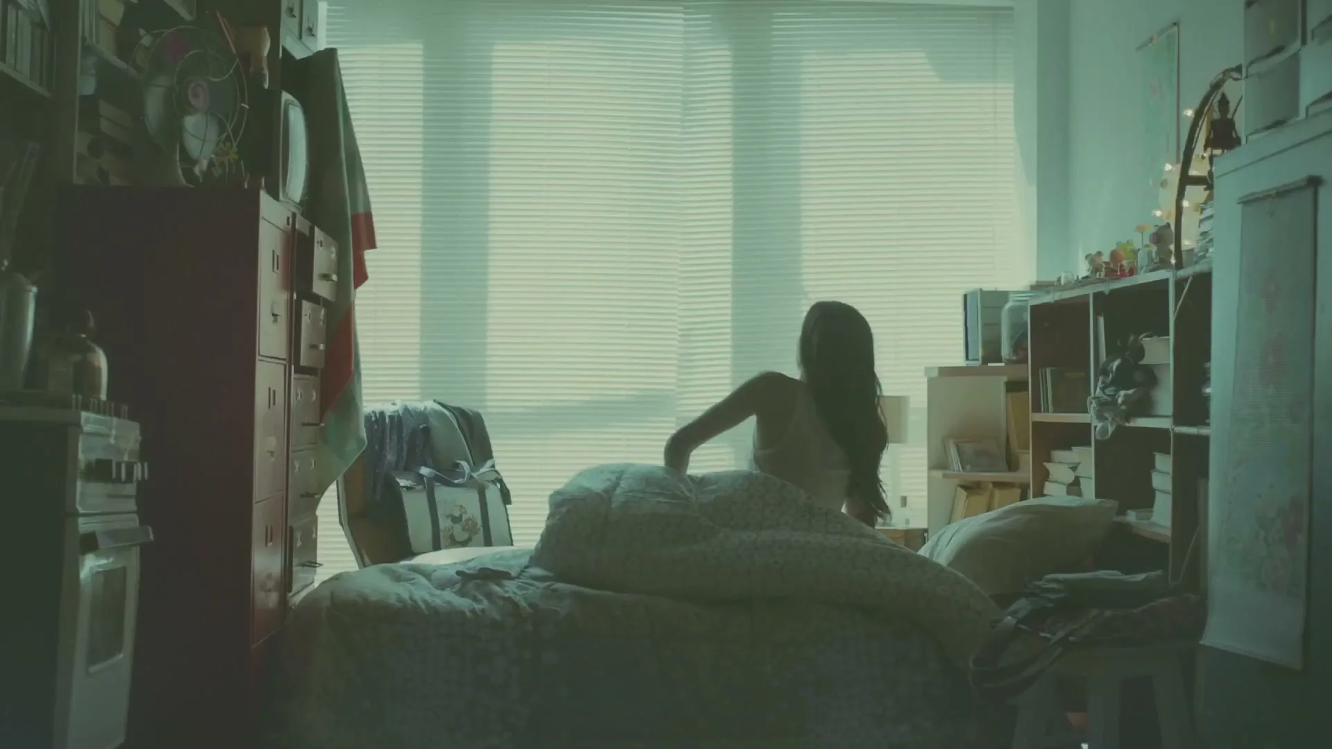 A young woman with long dark hair wakes up in a messy bedroom, sitting up in bed with a bright window behind her.
