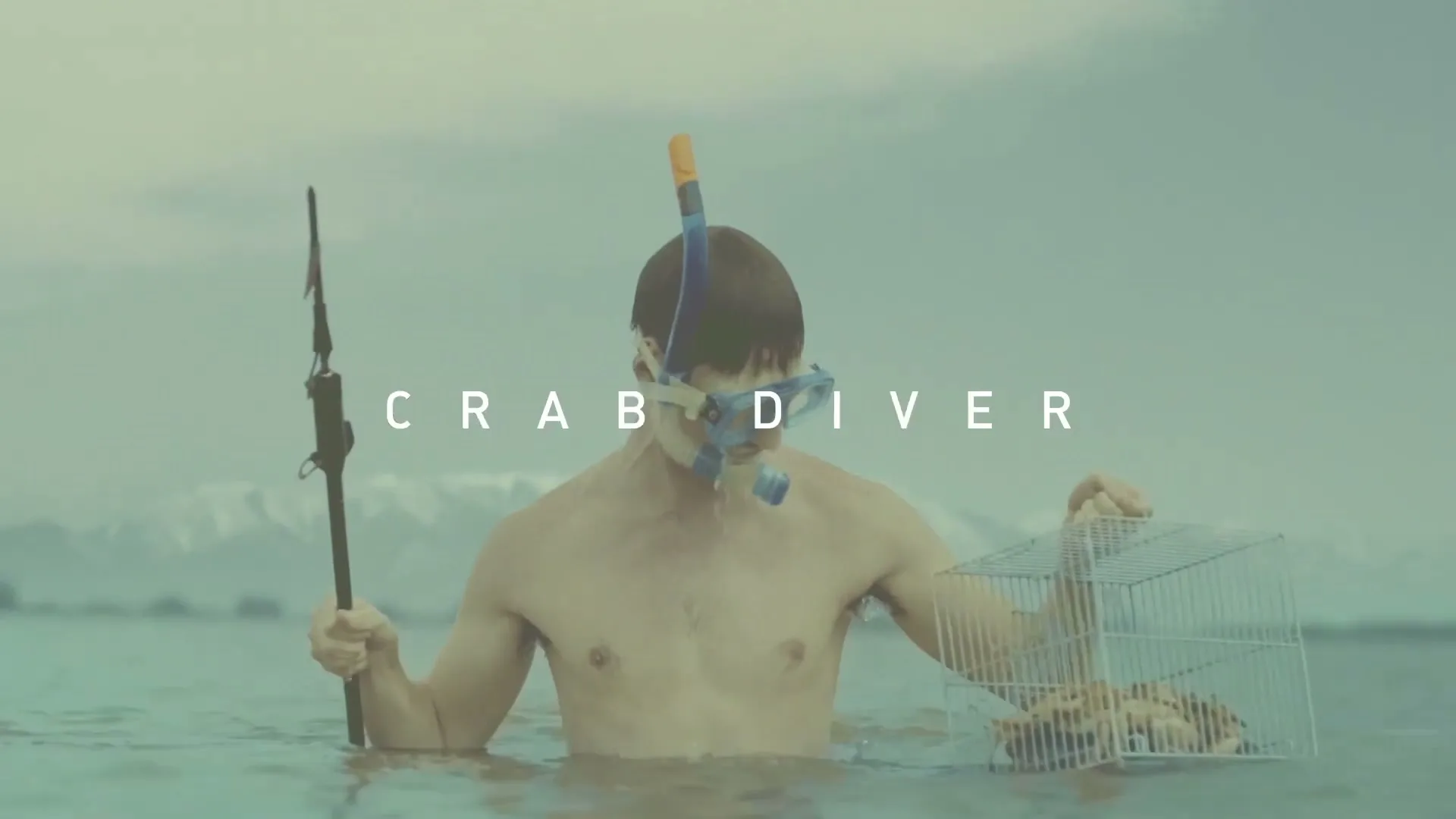 A man wearing a snorkel mask and holding a spear and a crab trap stands in the ocean with mountains in the background.