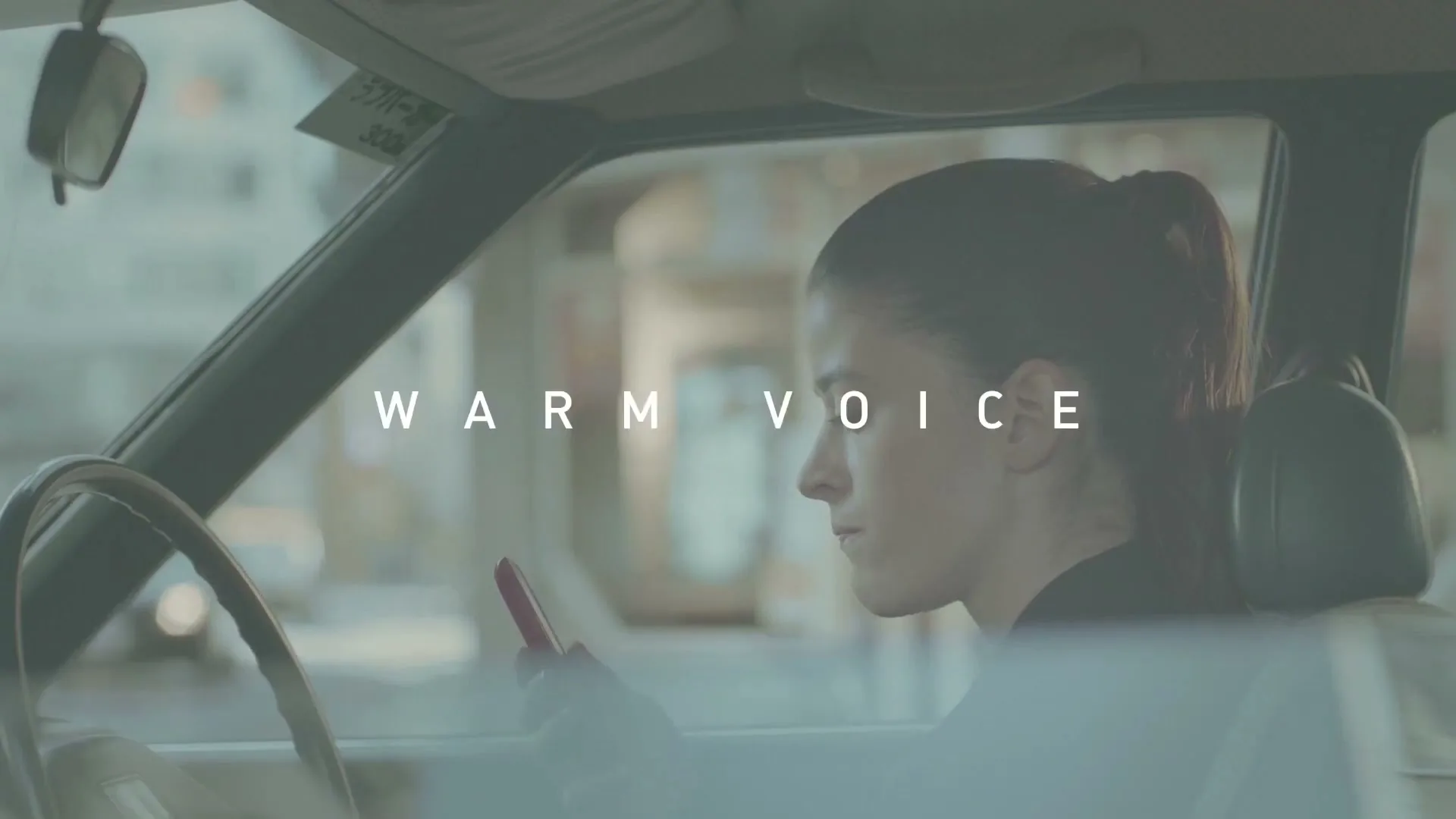 A young woman with dark hair in a ponytail sits in the driver's seat of a car, looking at her phone, with text appearing on screen.
