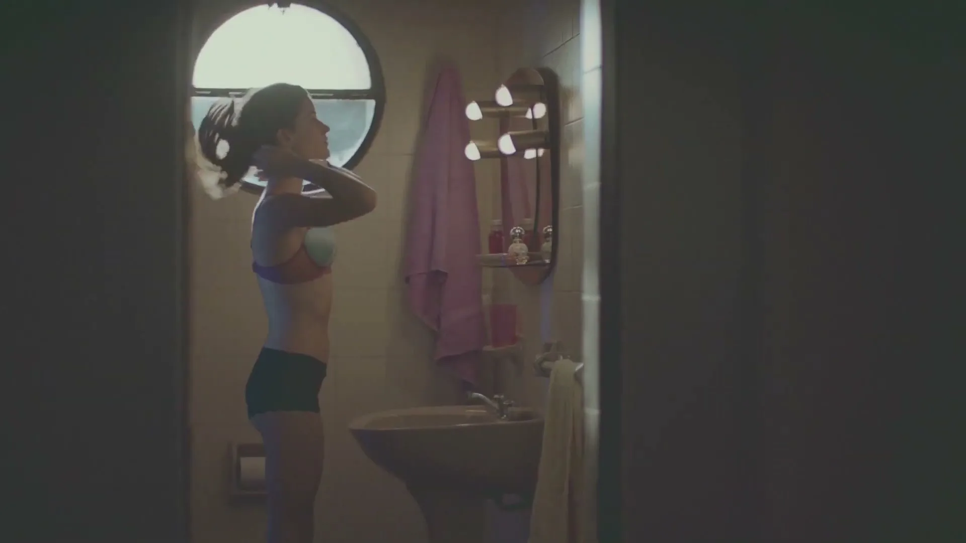 A young woman in her 20s, wearing a bra and shorts, stands in a bathroom, adjusting her hair in front of a mirror.