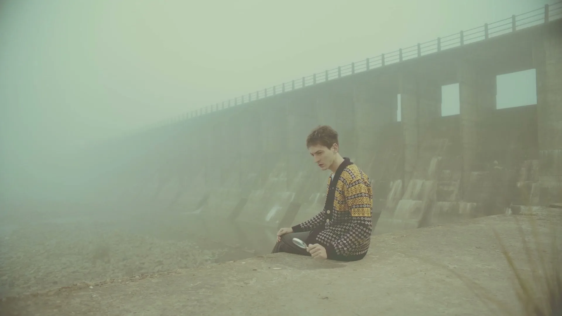 A young man in a patterned cardigan sits on a concrete ledge, looking down at a magnifying glass in his hand, with a large dam shrouded in mist behind him.