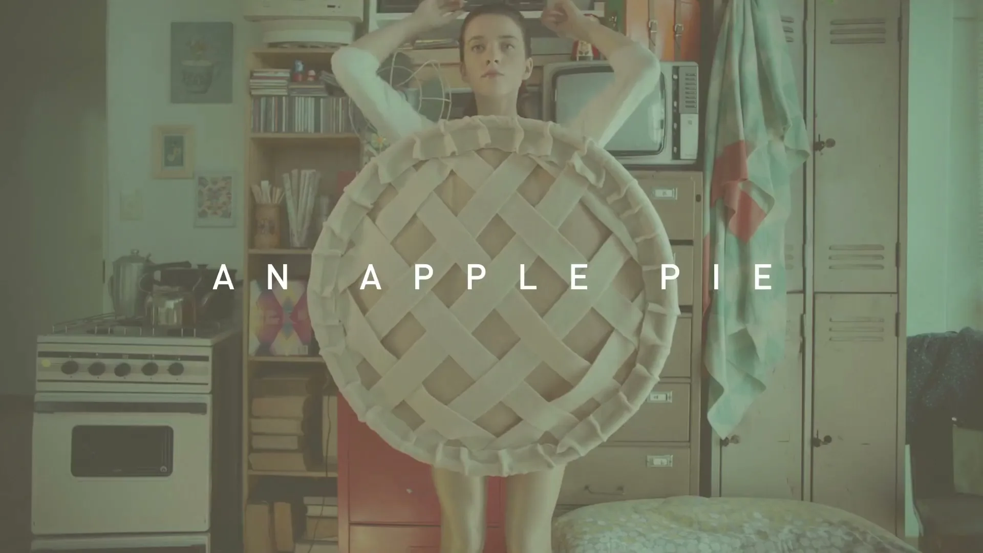 A young woman in a white top and beige shorts, wearing a large pie crust costume, stands in a cluttered room with text on screen.