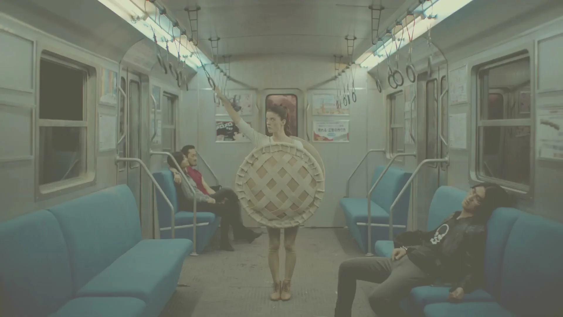 A woman in a pie costume stands in a subway car, holding onto a strap, while other passengers sit around her.