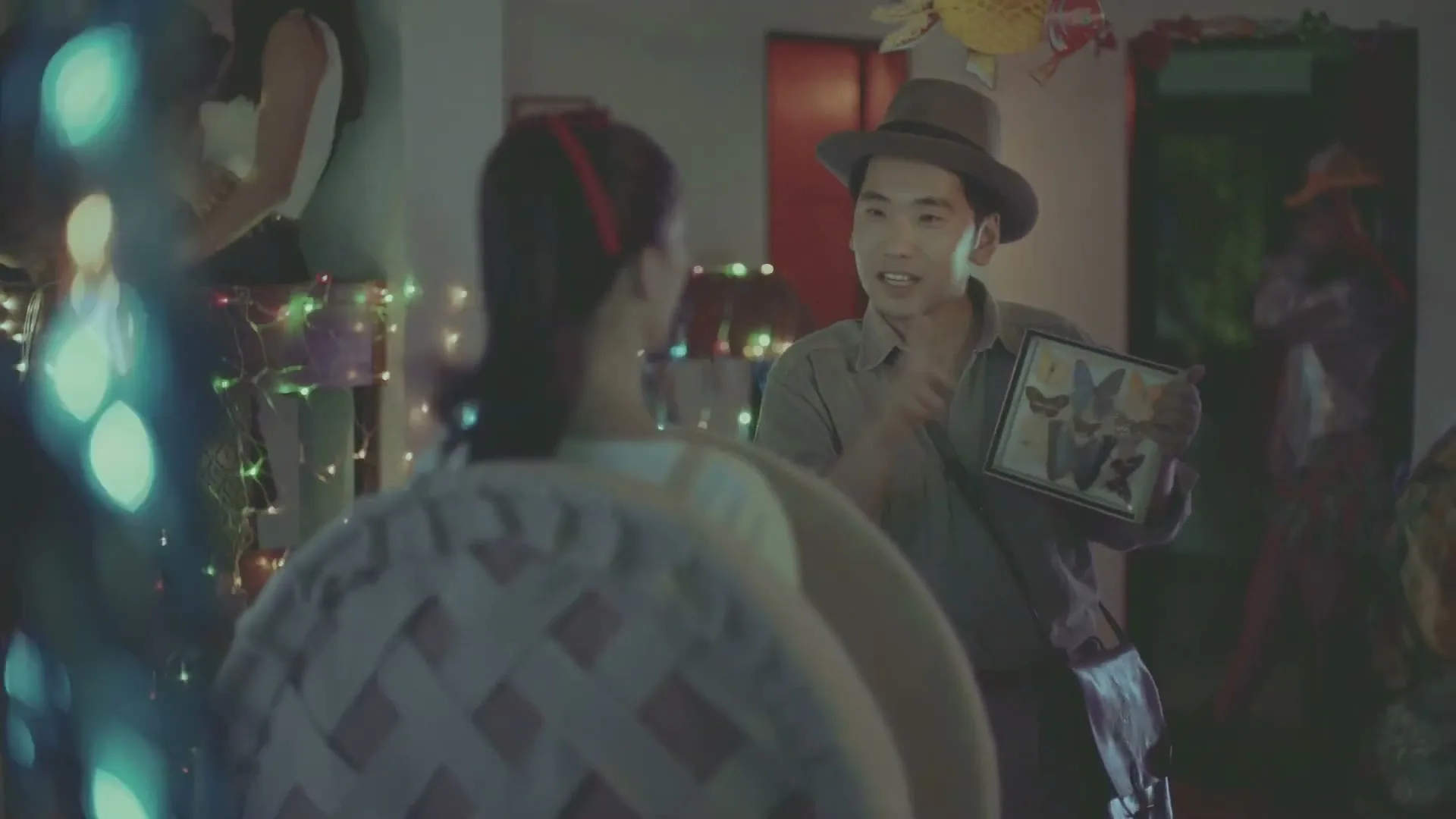 A man in a hat holds up a framed picture of butterflies to a woman, gesturing with his other hand at a festive party.