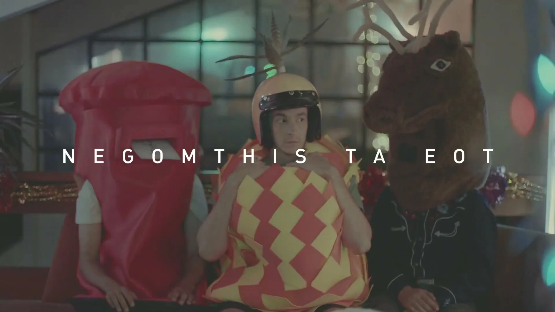 Three people in costumes sit on a couch, as one in a yellow and red checkered costume reacts to a pizza being offered, in a living room.