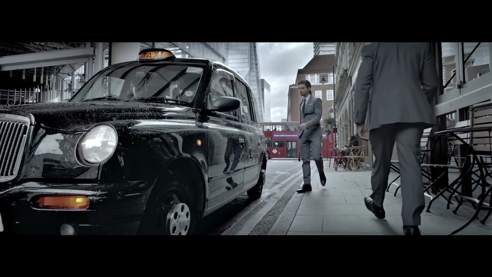 A man in a grey suit walks past a black taxi cab on a city street, with another man walking away in the foreground.