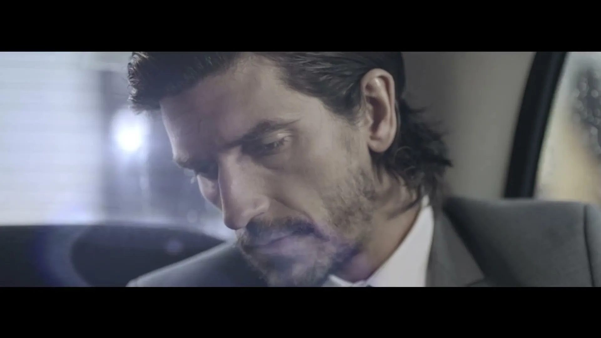 A man with dark hair and a beard, wearing a suit, looks down thoughtfully in the back of a car on a rainy day.