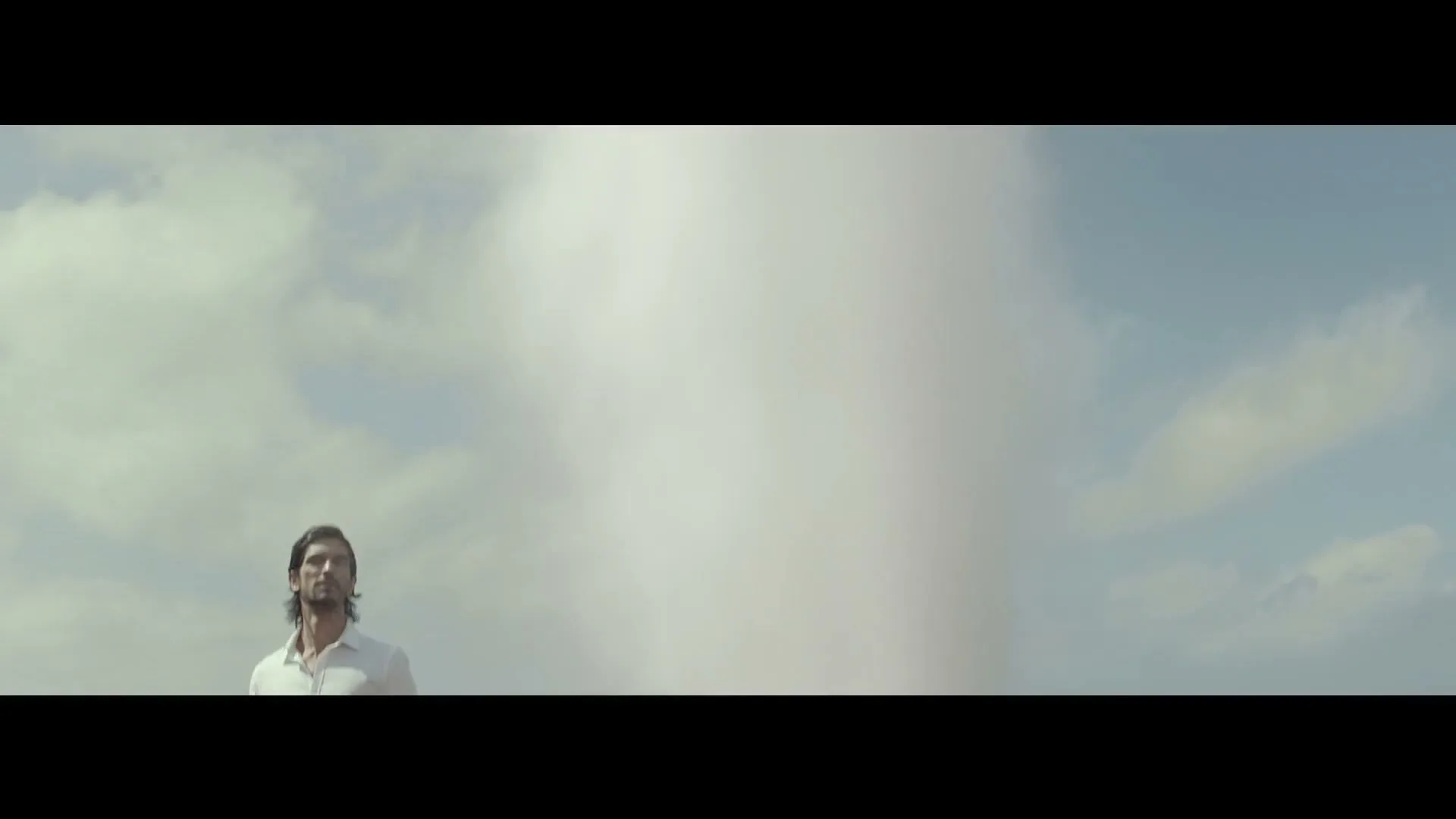 A man in a white shirt stands looking up at a large geyser of water against a cloudy sky.