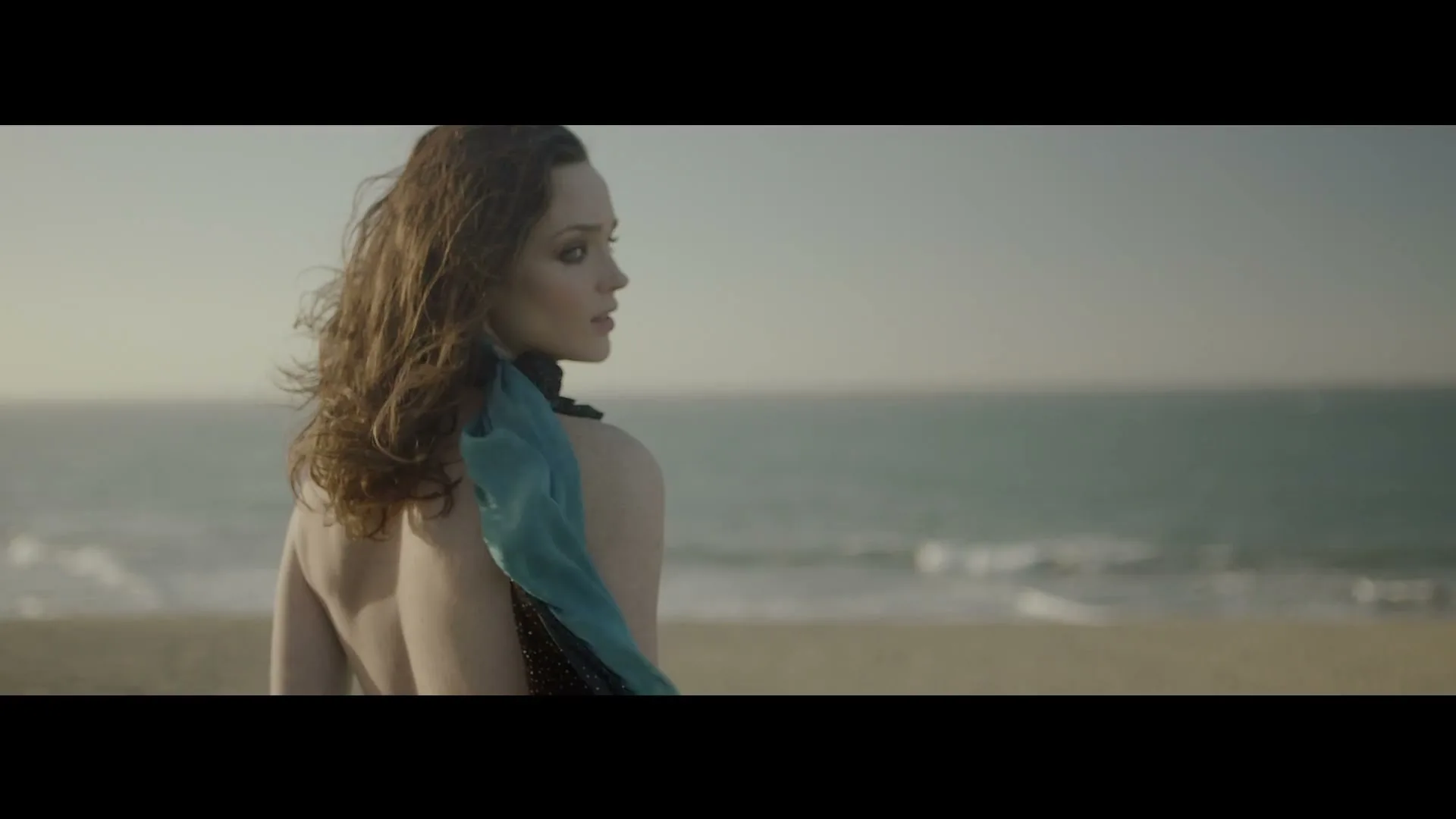 A woman with long, wavy brown hair and a blue scarf looks over her shoulder at the ocean on a sandy beach.