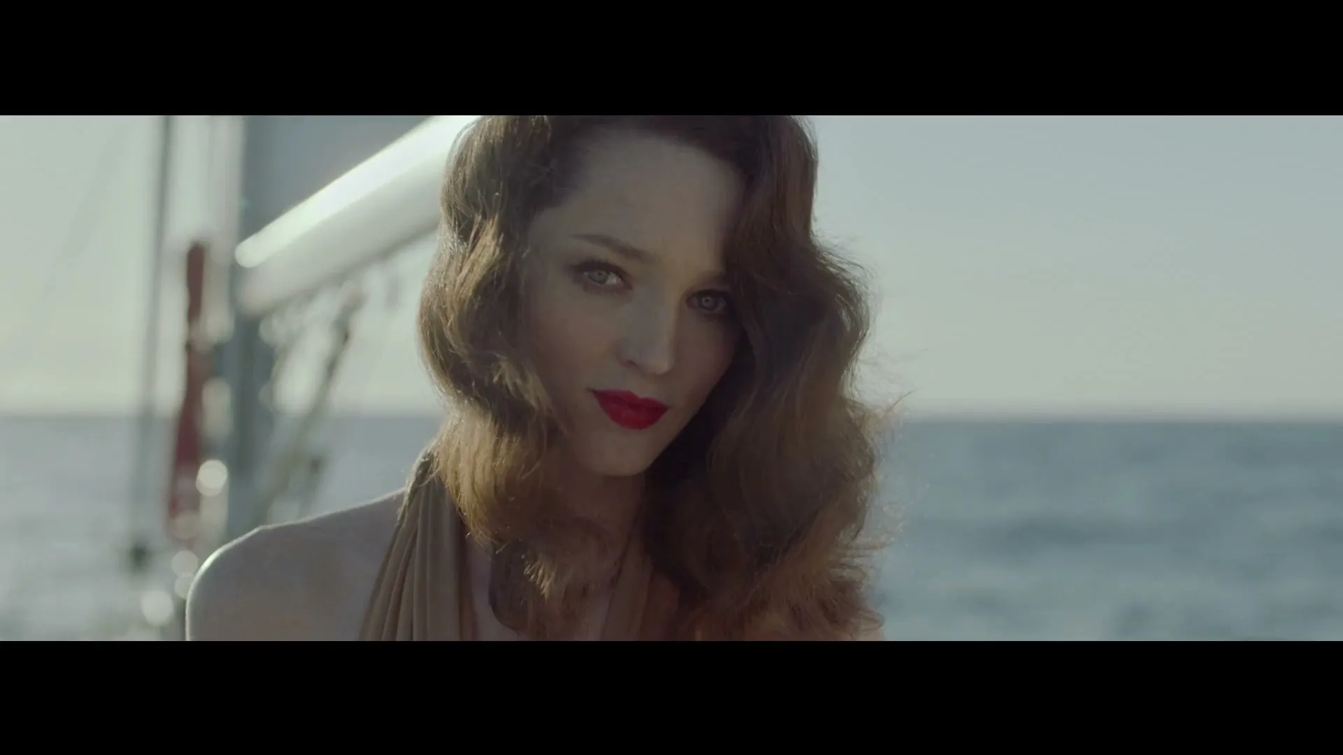 A woman with red lipstick and wavy hair looks directly at the camera, standing on a boat with the ocean in the background.