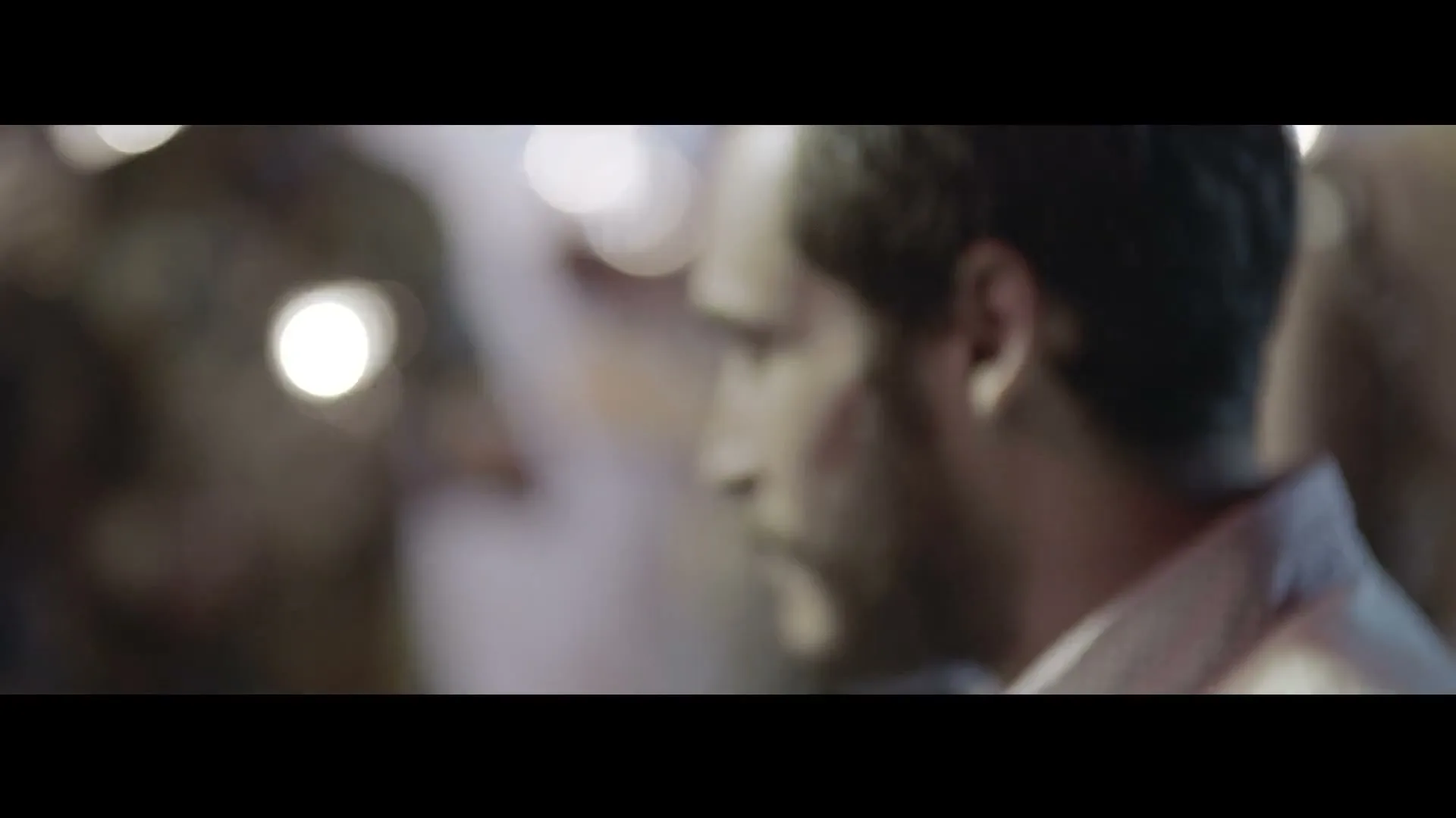 A man with a beard, wearing a collared shirt, is seen in profile, looking left, in a blurry, crowded interior setting.