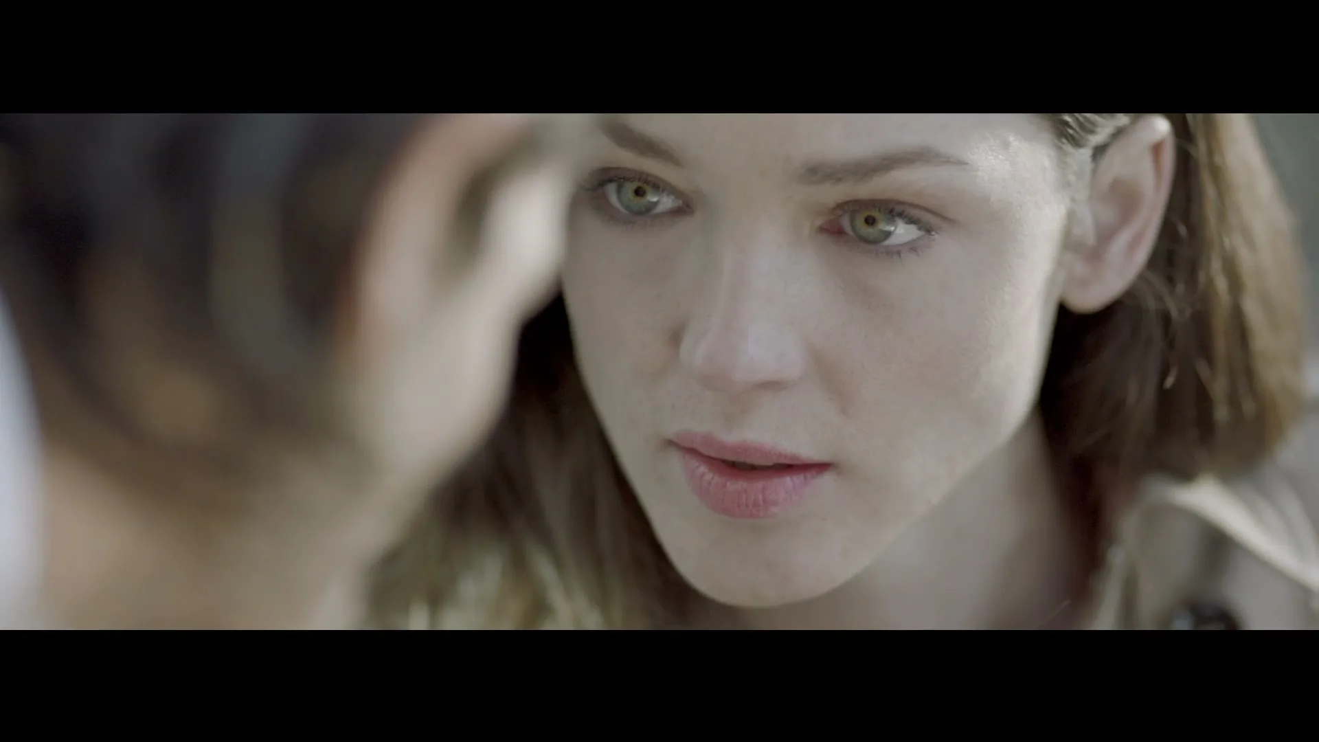 A young woman with green eyes and light brown hair looks intently at someone off-screen, her expression serious and focused.