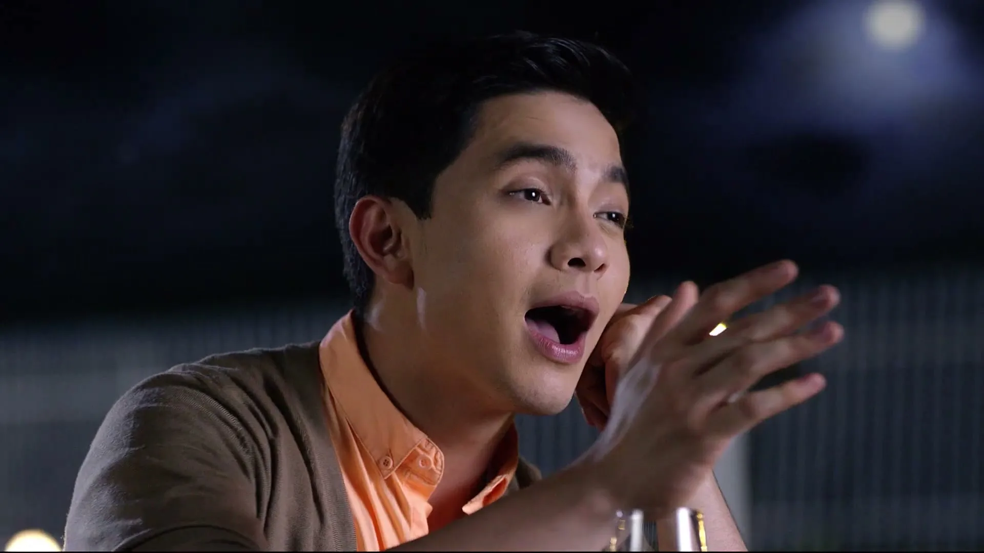 A young man in a peach shirt and brown cardigan, with dark hair, is seen from the chest up, talking and gesturing with his hands in a dimly lit interior.