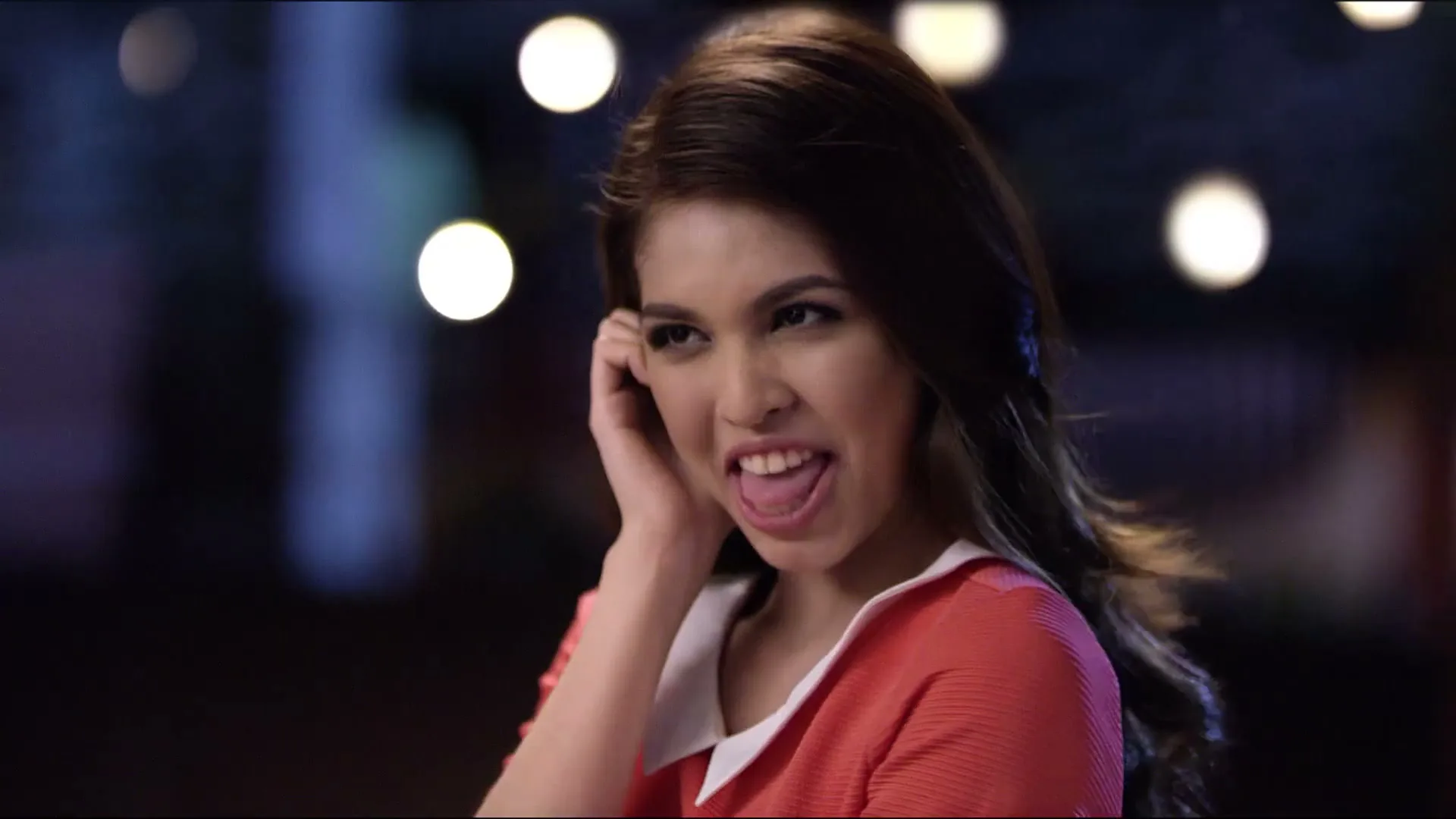 A young woman with long dark hair, wearing a red dress with a white collar, smiles and then makes a funny face, holding her hand to her ear, in a dimly lit outdoor setting at night.