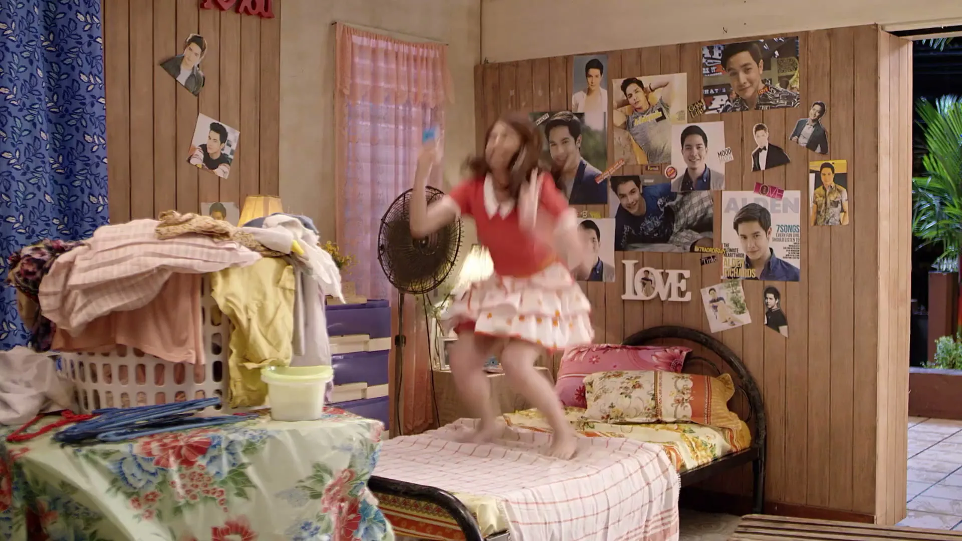 A young woman in a red dress and white apron jumps on a bed in a messy bedroom, holding a phone and smiling joyfully.