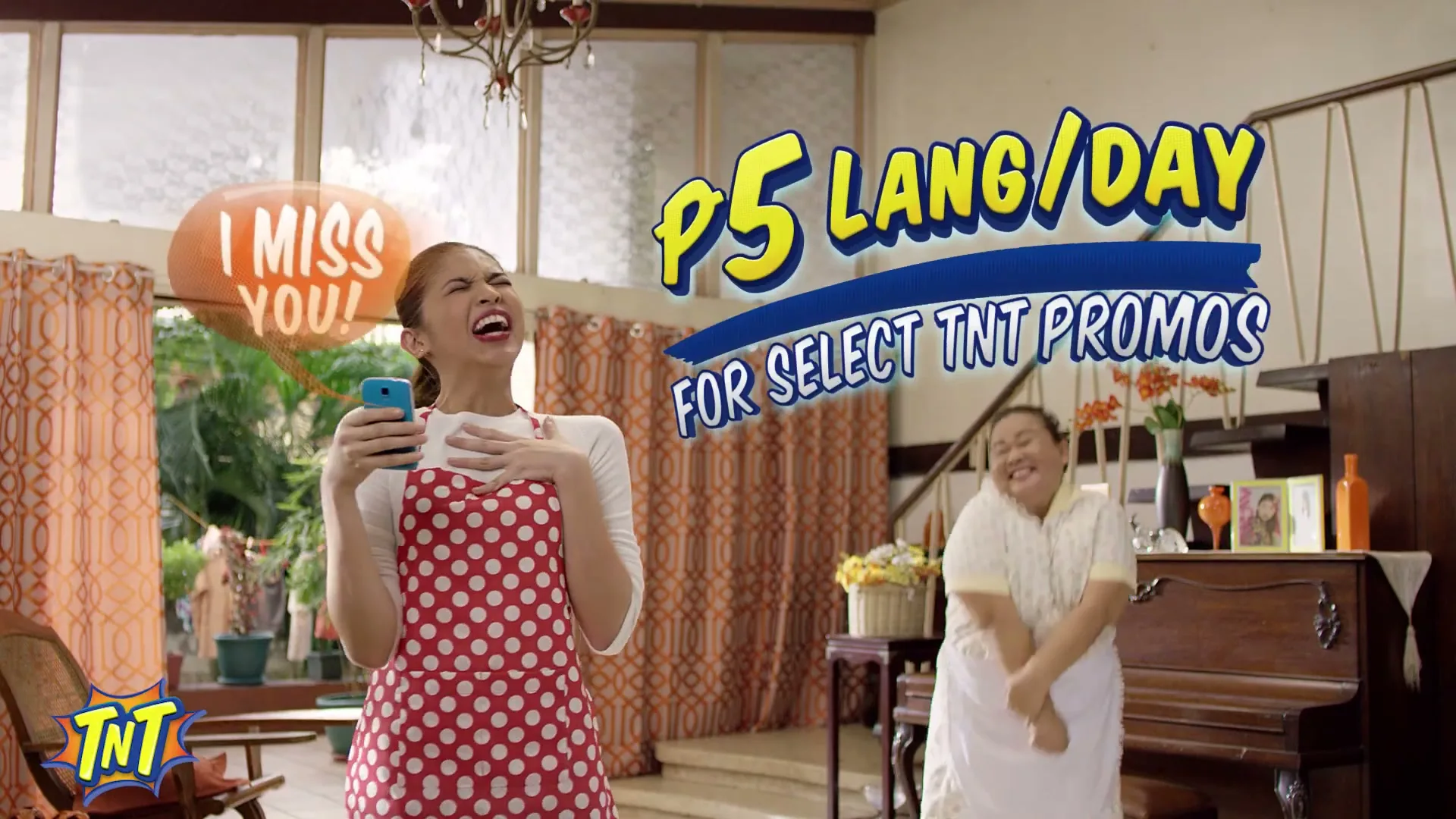 A young woman in a red polka dot apron laughs while looking at her phone, as an older woman in a white dress dances excitedly in a living room.