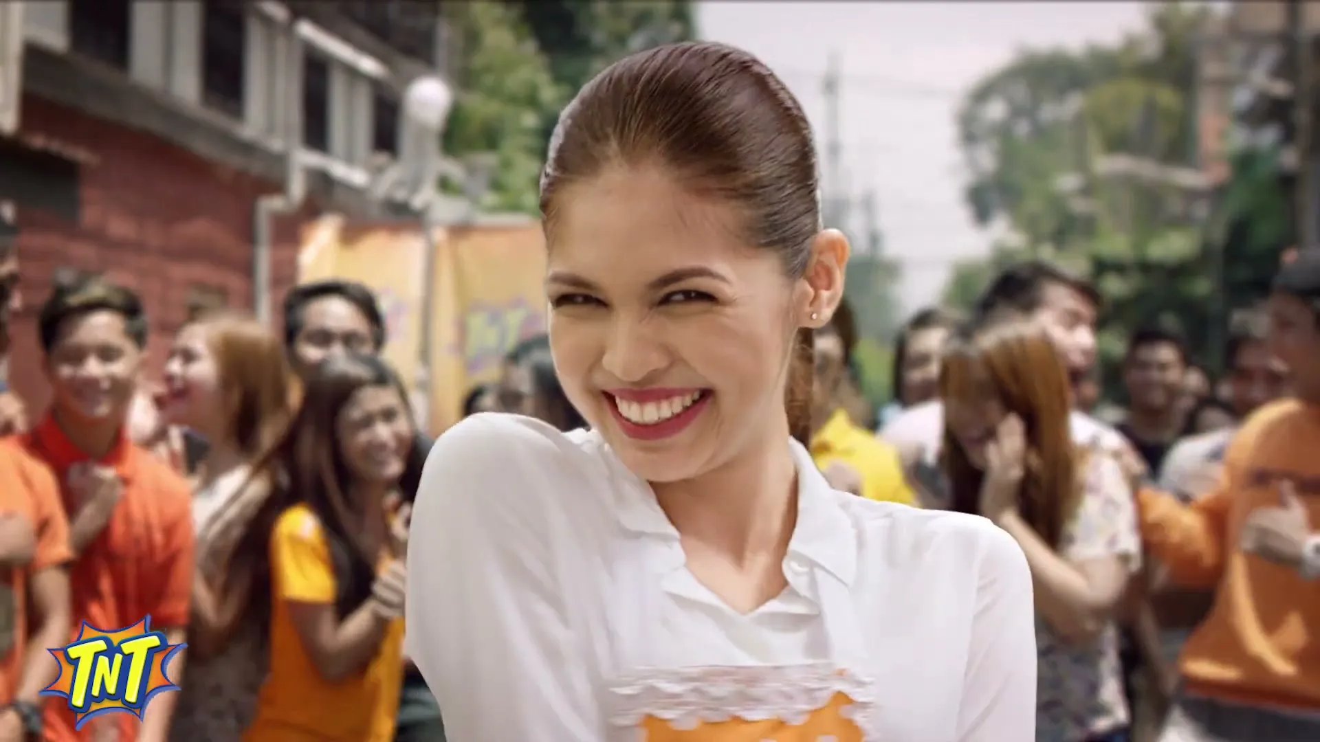 A young woman with a high ponytail, wearing a white shirt and an orange apron, smiles and winks playfully at the camera amidst a crowd of people on a sunny street.