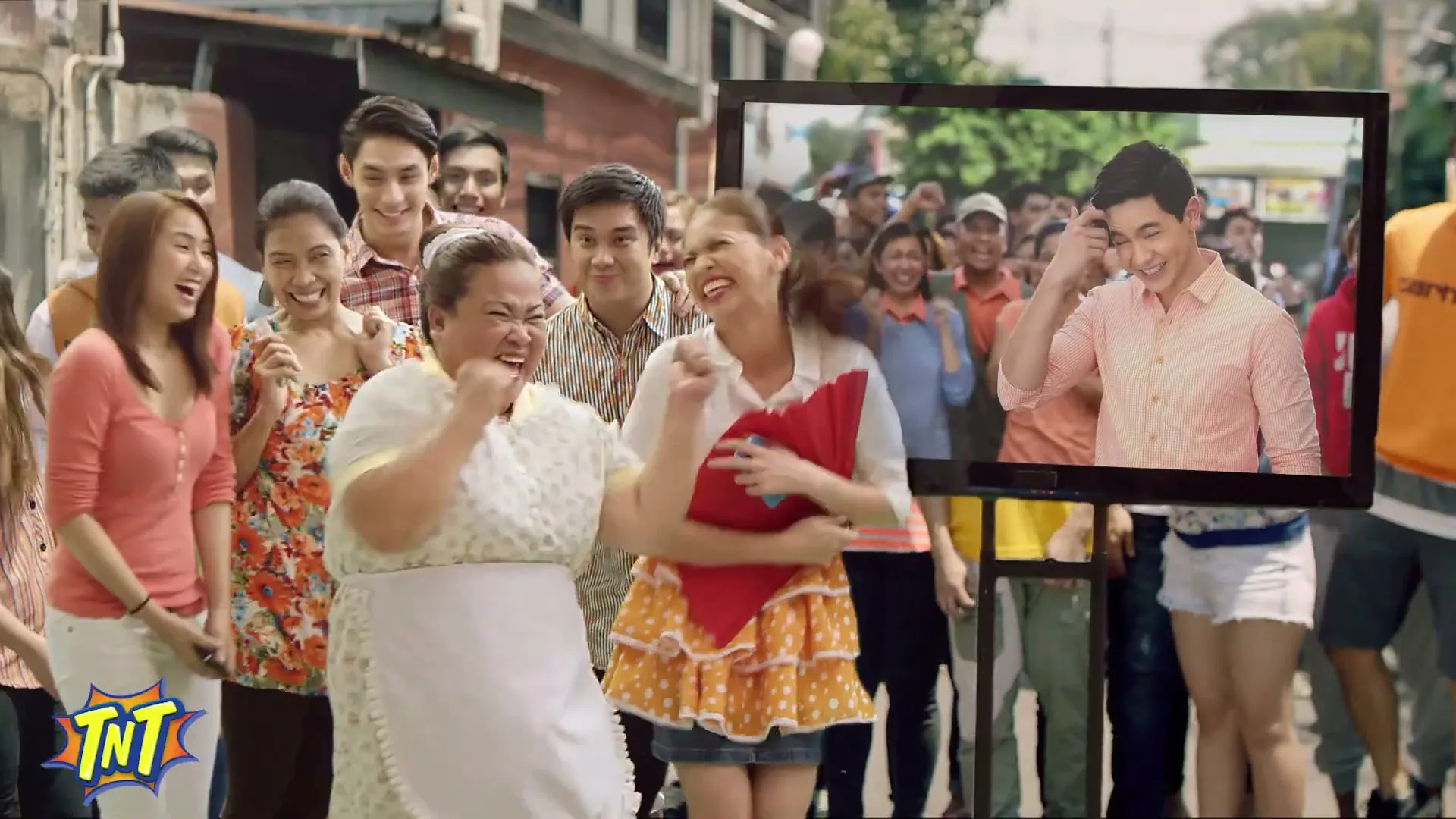 A diverse group of people, including a woman in an apron and a young woman in a polka dot skirt, cheer and dance around a TV screen showing a smiling man, on a sunny street.