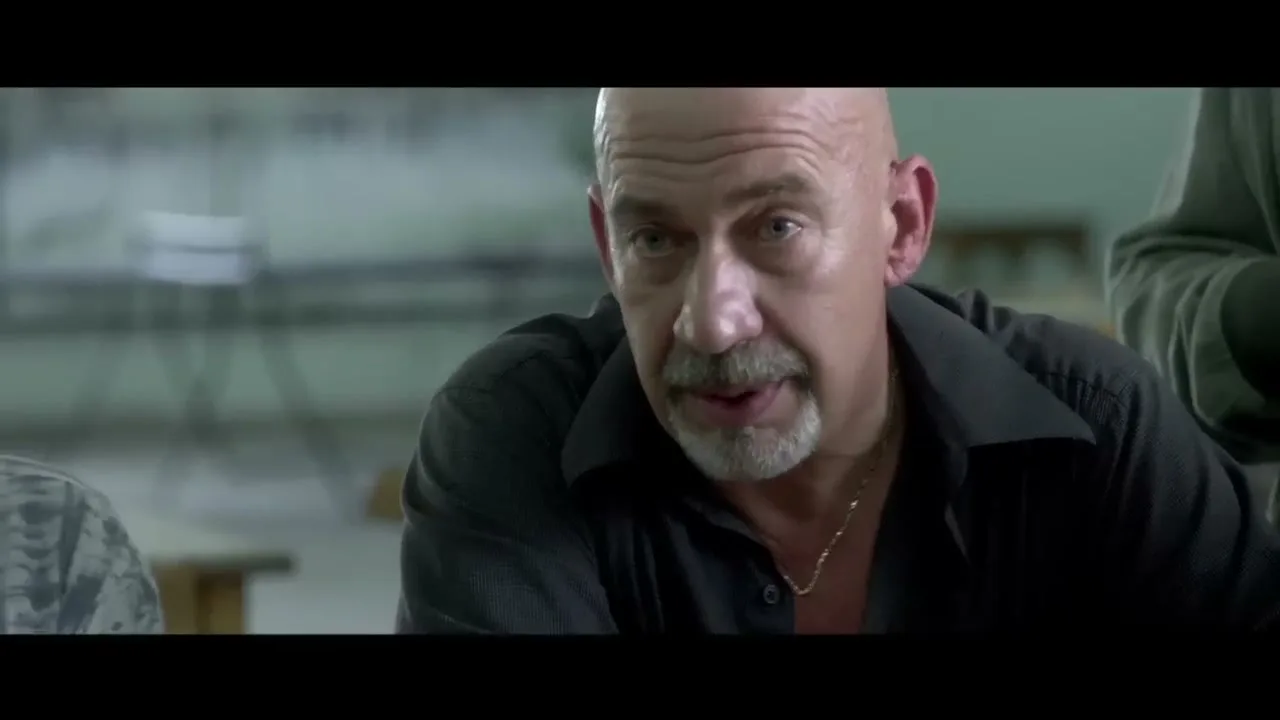 A bald, older man with a goatee and a gold chain talks to someone off-screen in a dimly lit room.