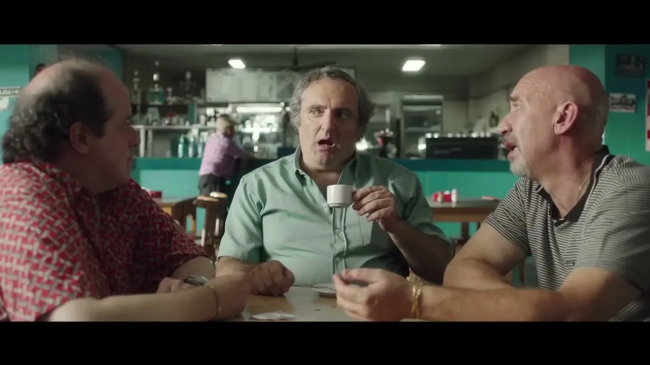 Three middle-aged men sit at a table in a diner, one holding a small cup, as they talk and listen intently to each other.