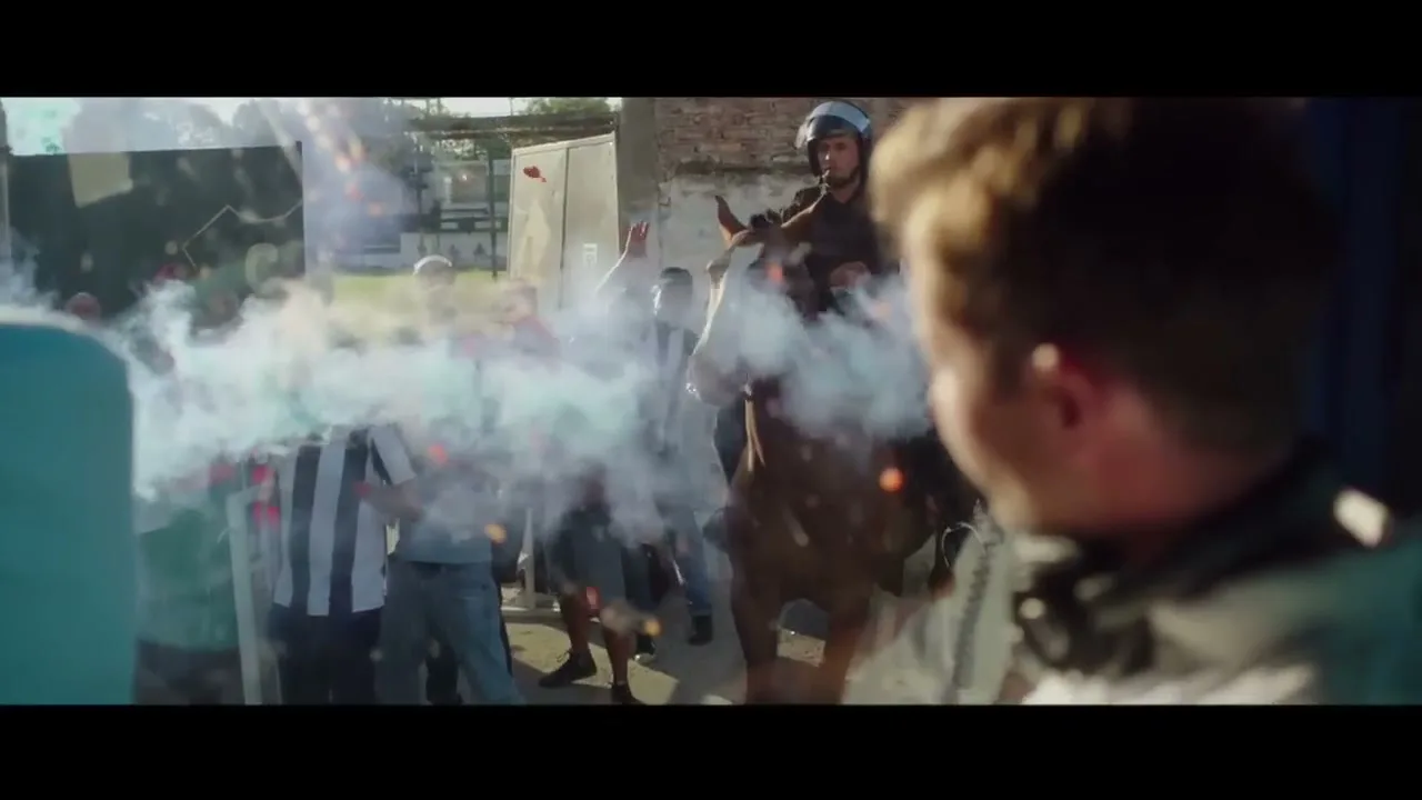 A man in a helmet on horseback rides through a crowd of cheering soccer fans, as smoke and confetti explode around them in an outdoor stadium.