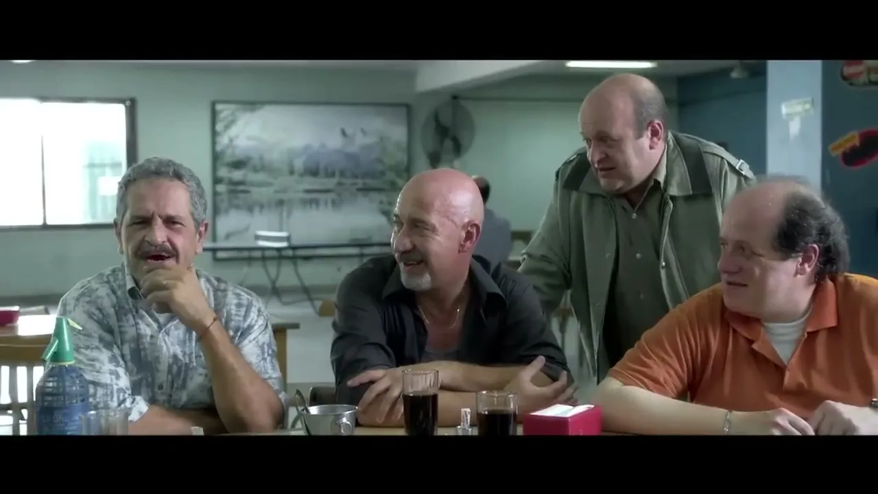 Four middle-aged men, three seated at a table and one standing, are engaged in conversation in a brightly lit cafeteria.
