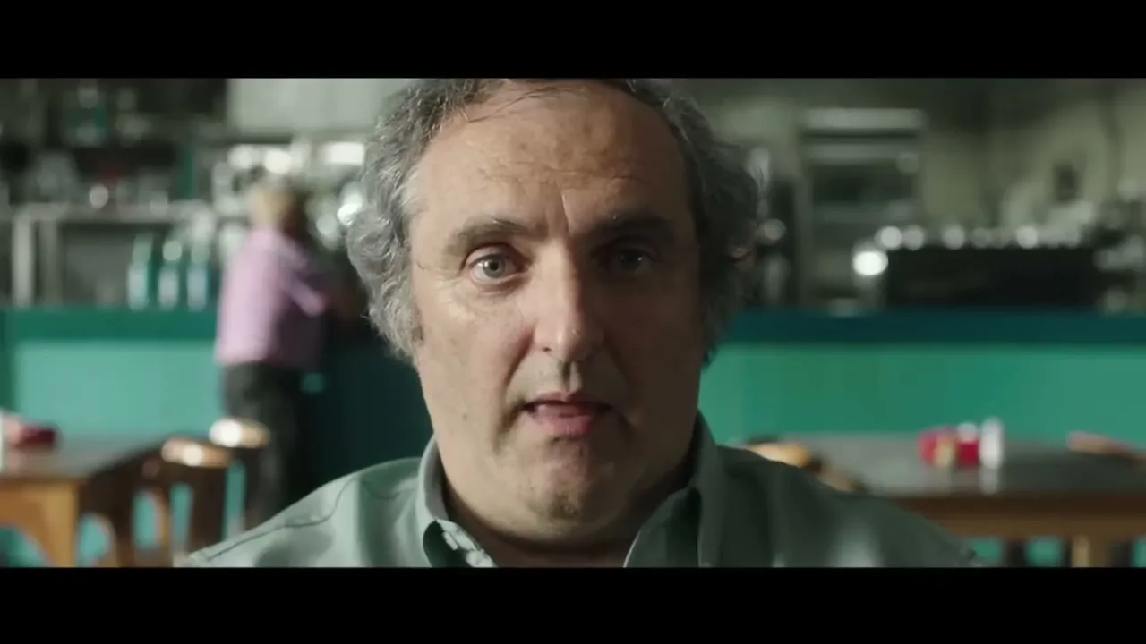 A middle-aged man with graying hair and a light blue shirt looks directly at the camera in a cafe, with a blurred background.