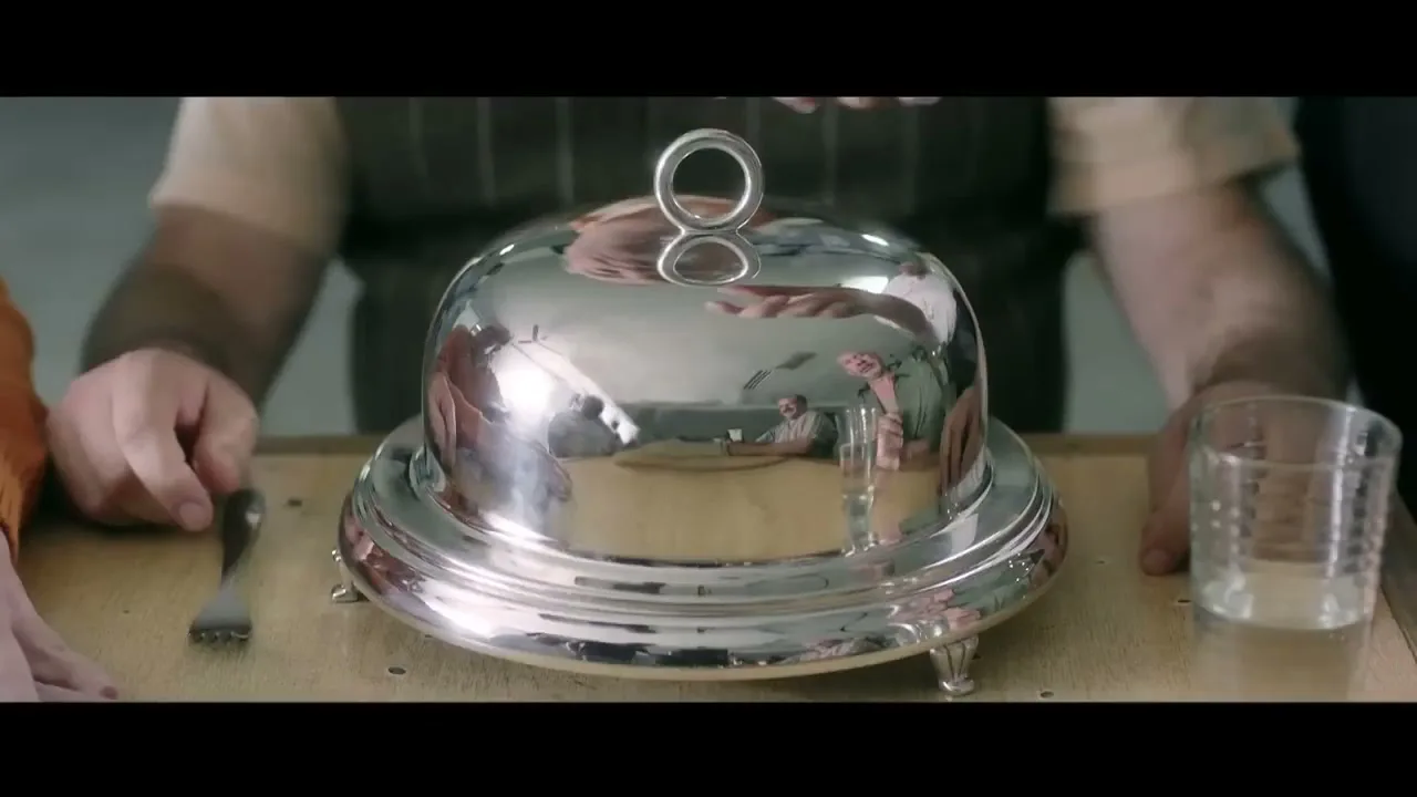 A waiter's hands are about to lift a silver cloche from a plate on a wooden table, with a glass and fork nearby.