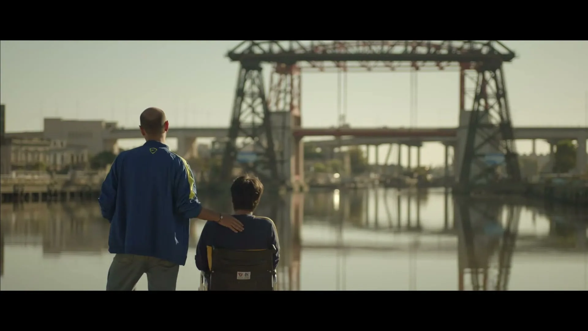 Two men, one standing and one in a wheelchair, look out over a river with a large industrial bridge in the background on a sunny day.