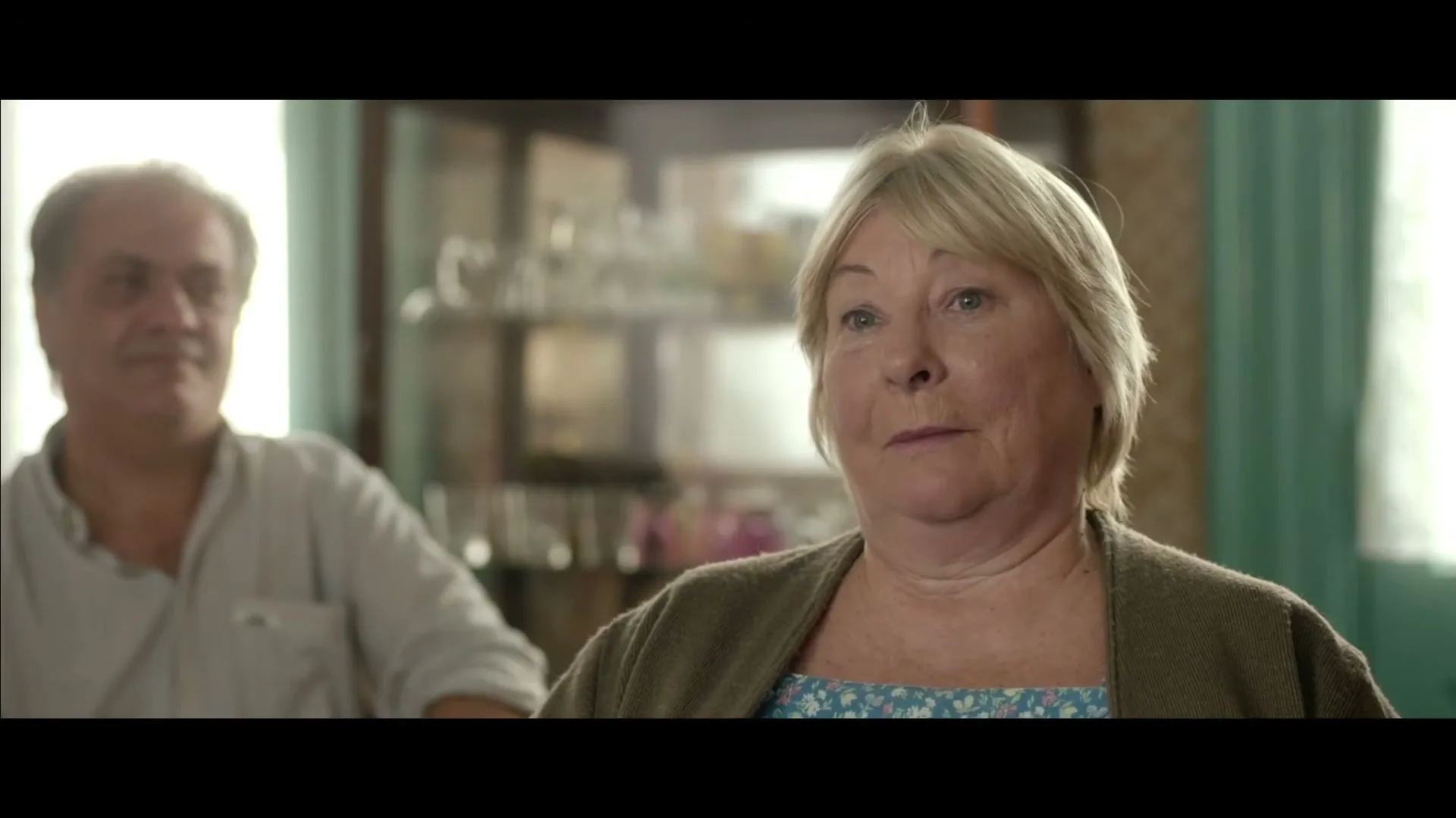 A blonde woman in her 60s, wearing a brown cardigan and blue floral top, speaks while a man in his 40s sits out of focus behind her in a sunlit room.