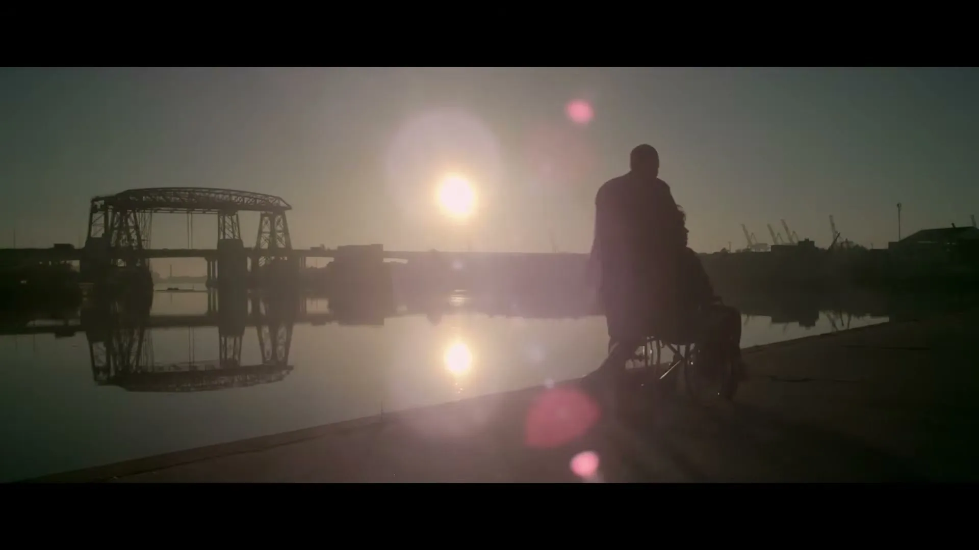 A man pushes another person in a wheelchair along a waterfront at sunset, with a bridge and industrial cranes in the background.