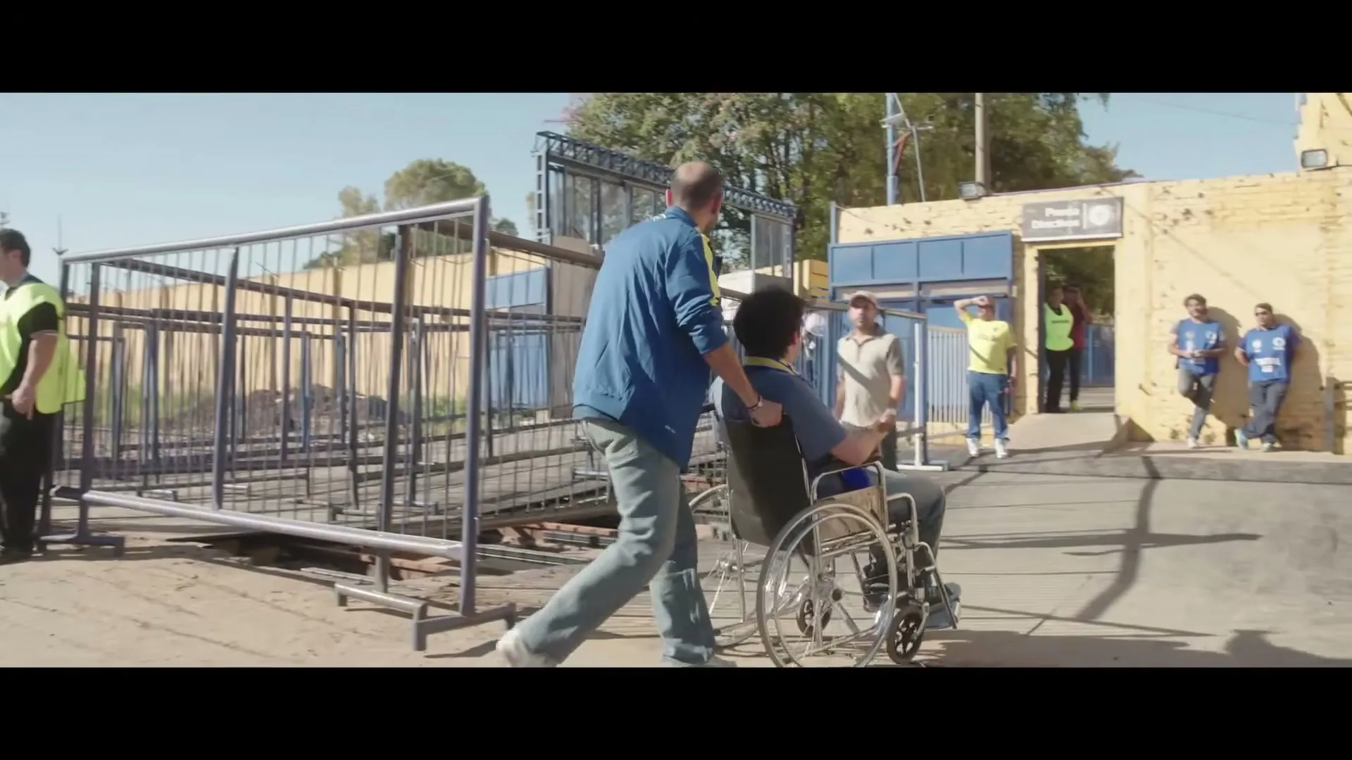 A man pushes another man in a wheelchair, who is wearing a blue and yellow scarf, towards a stadium entrance on a sunny day.