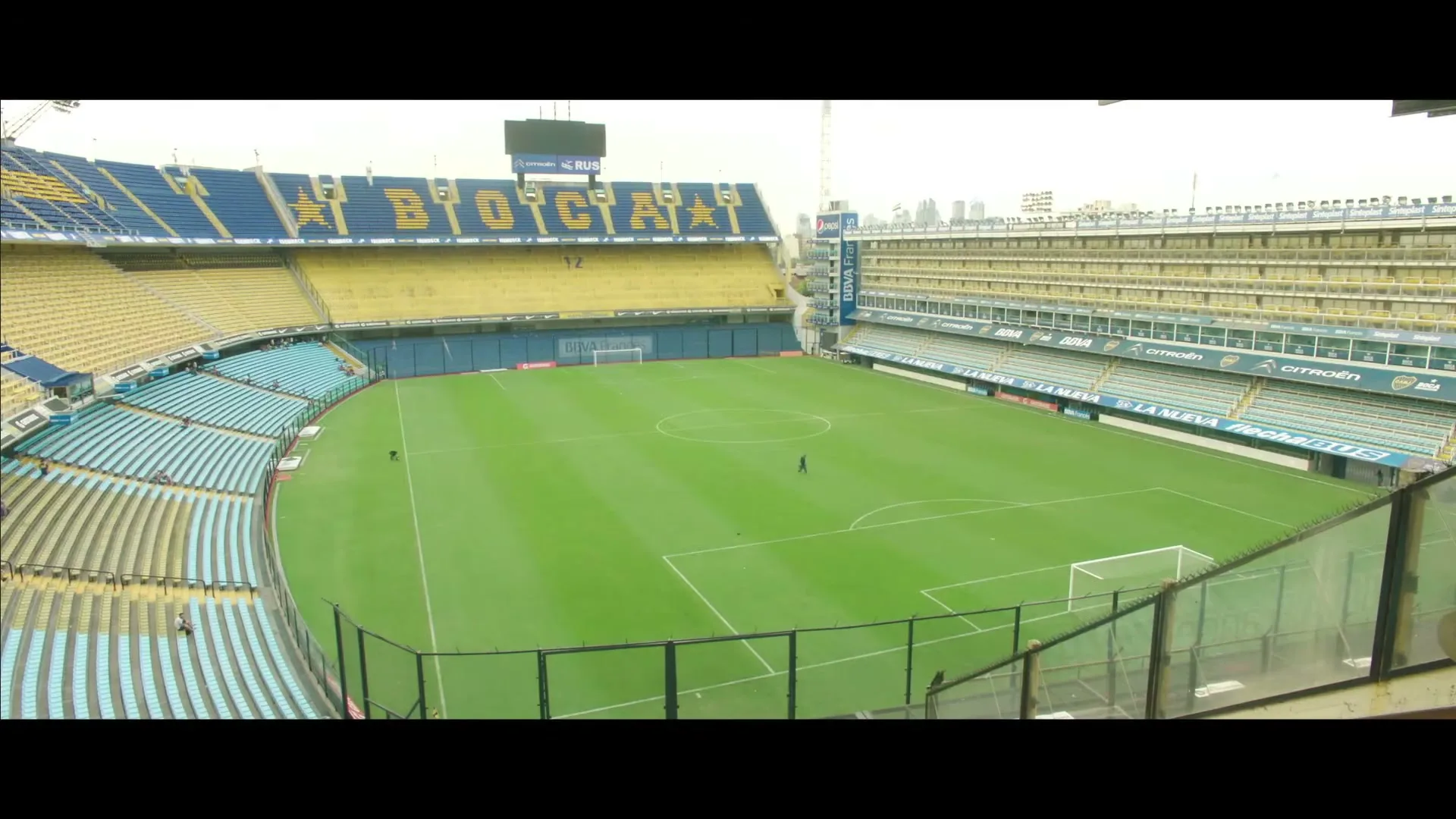 An empty soccer stadium with green grass and blue and yellow seats is shown from a high angle, with a few people walking on the field.