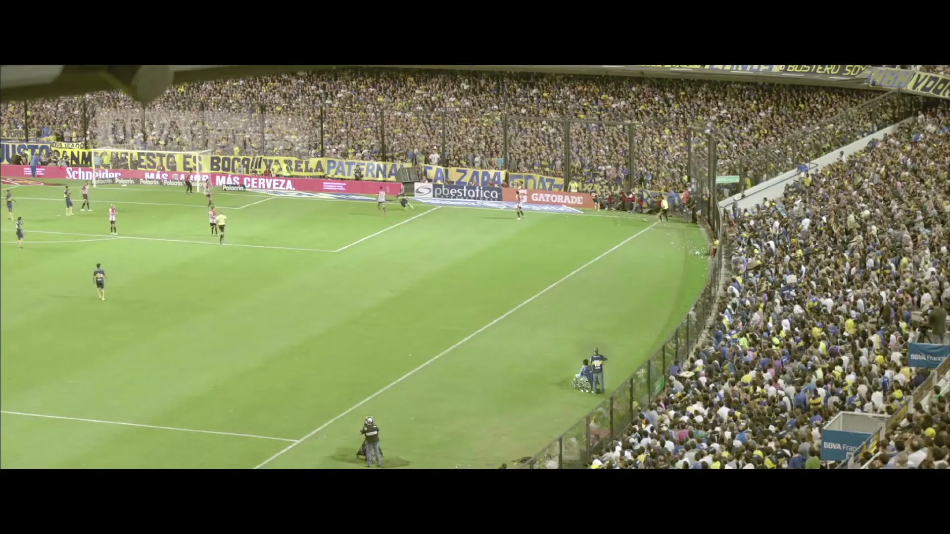 A wide shot from a high angle shows a soccer match in progress on a green field, surrounded by a massive crowd of cheering fans in a large stadium.