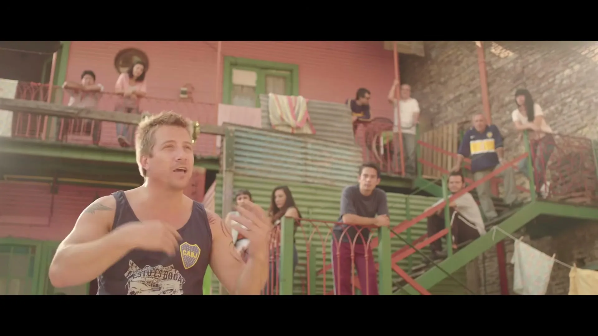 A man in a blue tank top with a tattoo on his arm talks to the camera, surrounded by people on balconies and stairs of colorful buildings in a vibrant neighborhood.