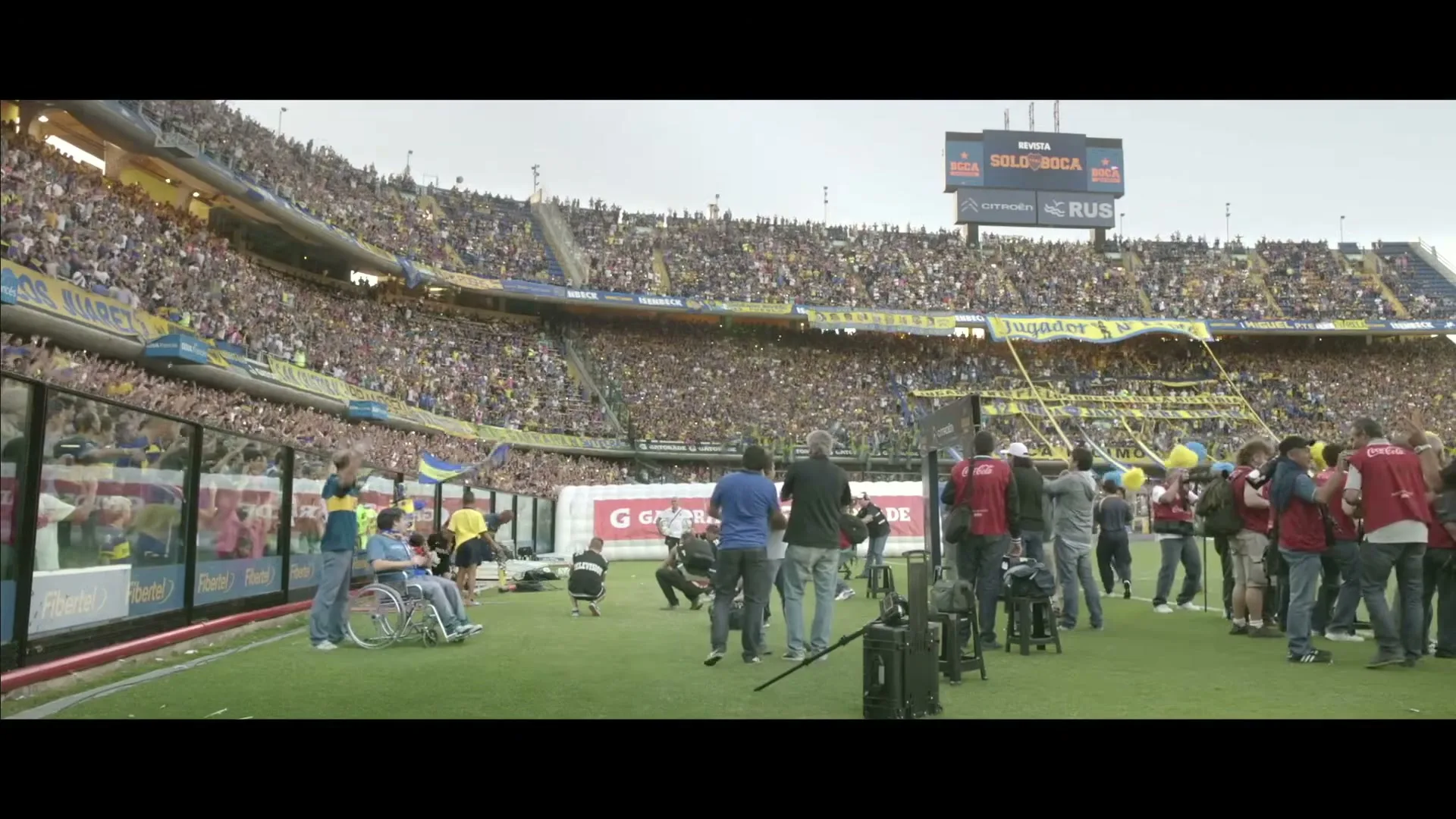 A wide shot shows a large crowd in a stadium, with a man in a wheelchair on the field, surrounded by crew members.