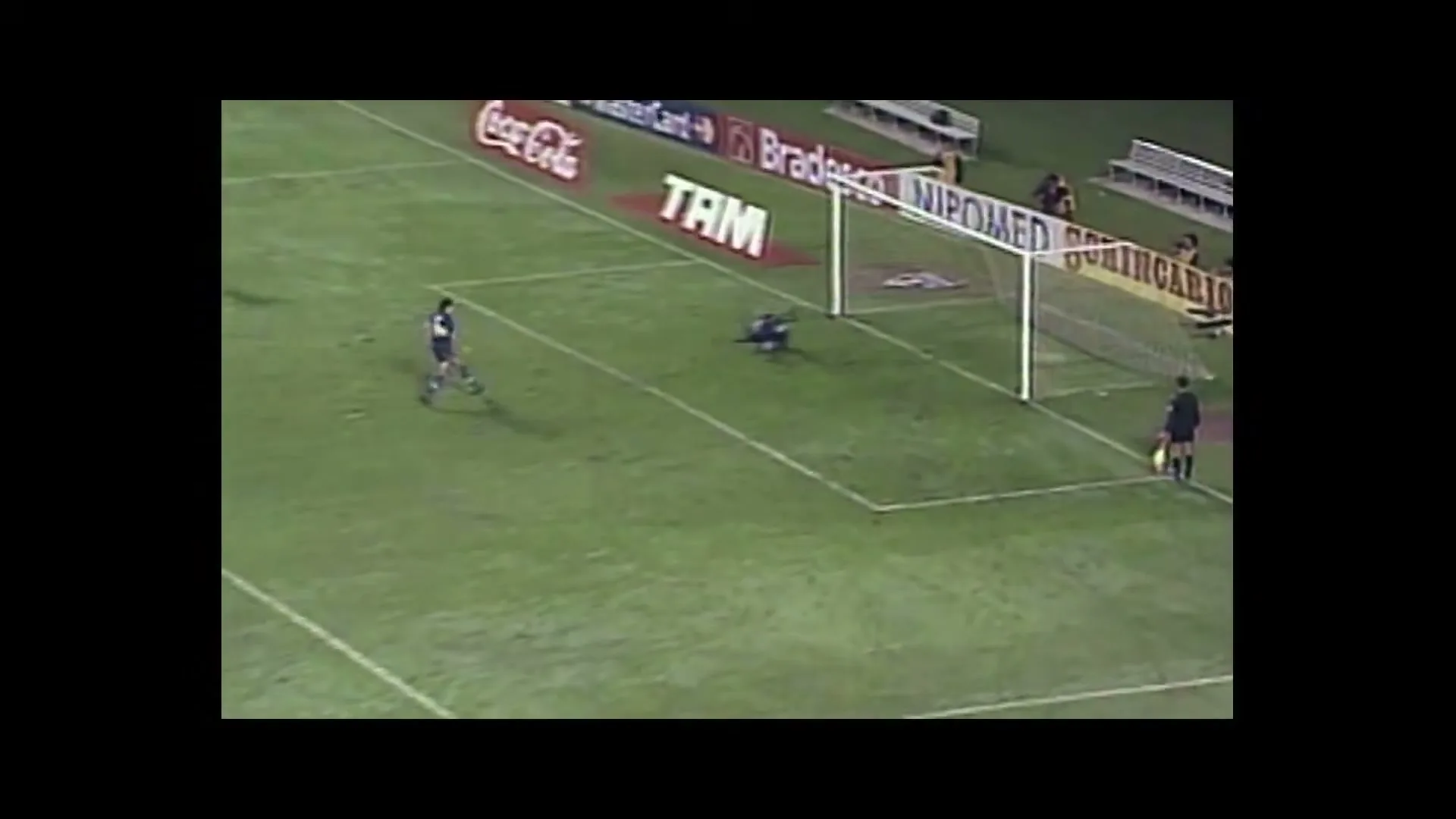 A soccer player in a blue and yellow uniform kicks a penalty shot towards the goal, while the goalkeeper dives to block it on a green field at night.