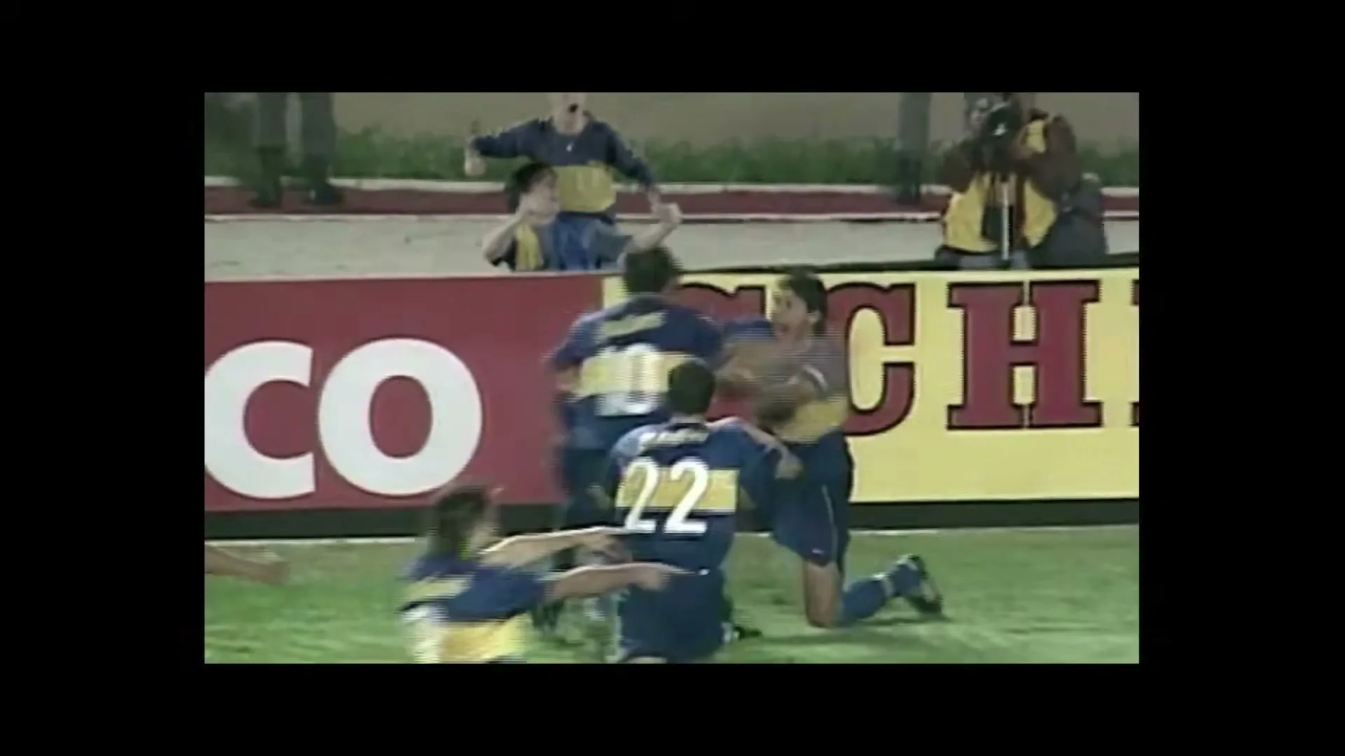 A male soccer player in a blue and yellow uniform slides on his knees in celebration, then is joined by his teammates on a green field with a red and yellow sign in the background.