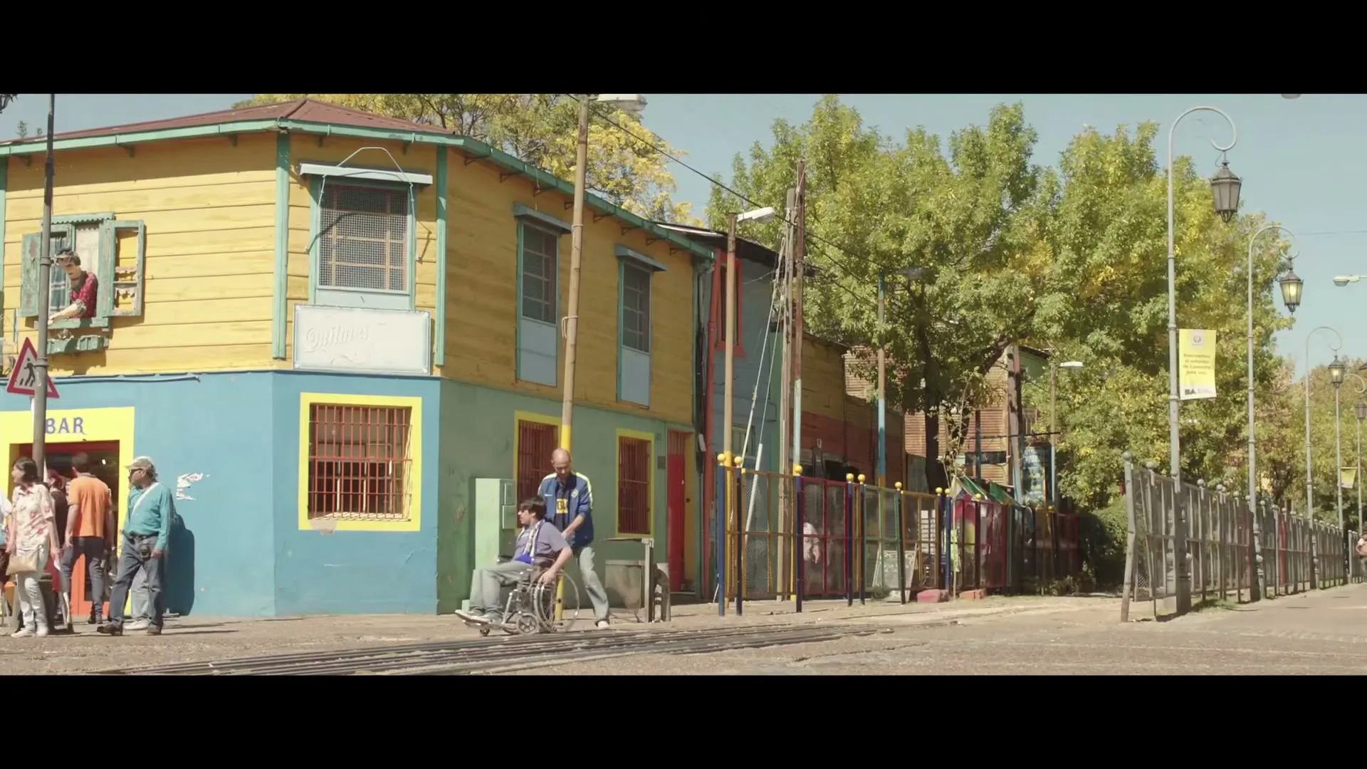 A man in a blue tracksuit pushes a man in a wheelchair down a street with colorful buildings and trees, as other people walk by.