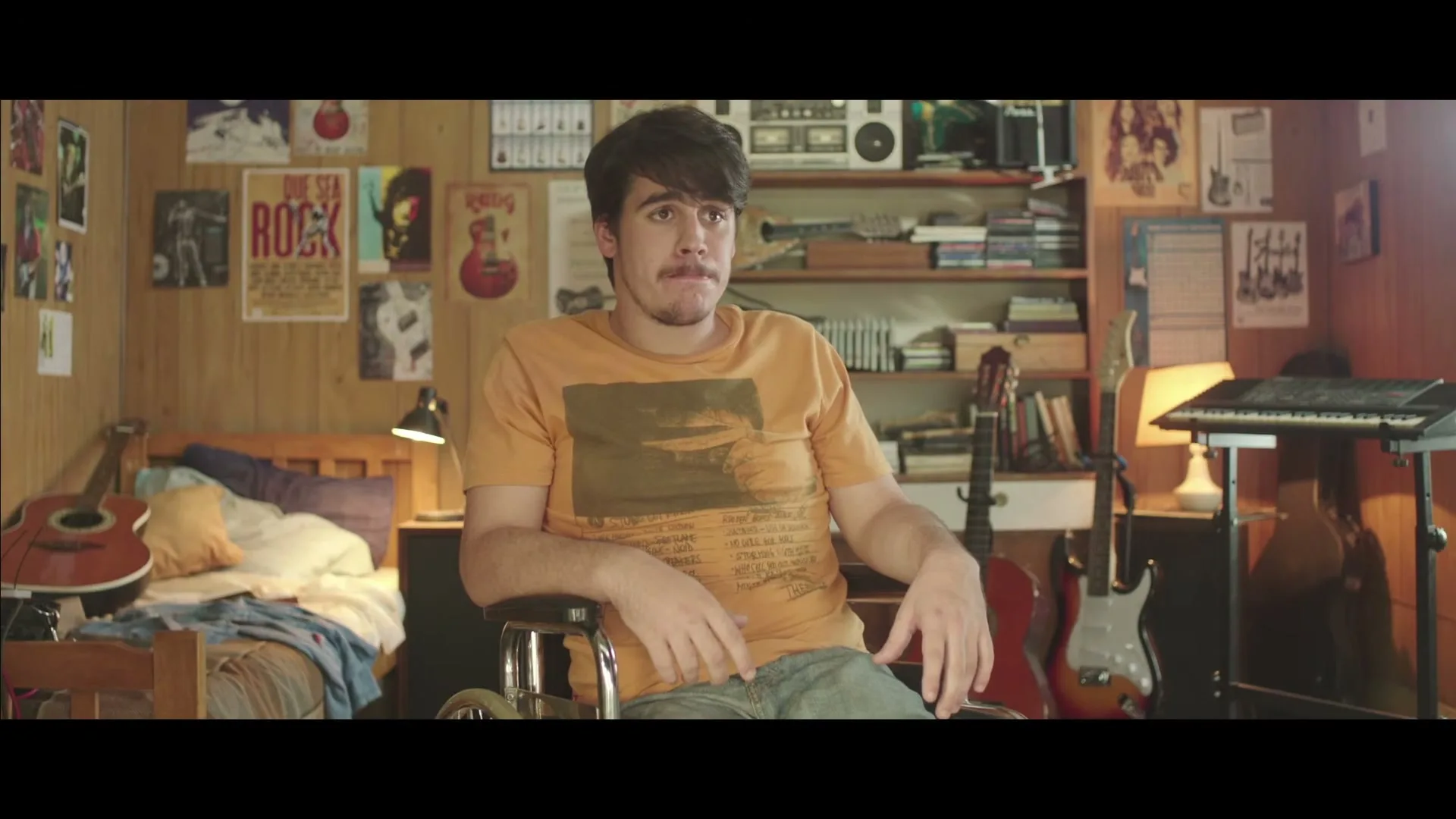 A young man with dark hair and a mustache sits in a wheelchair, wearing an orange t-shirt, looking surprised in his bedroom.