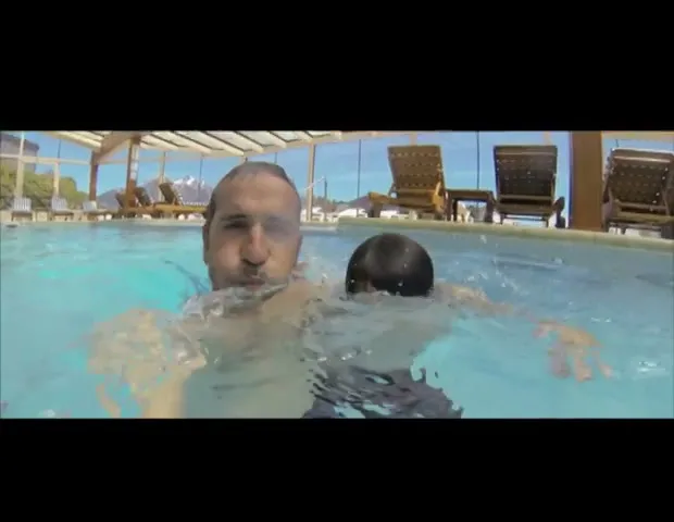 A father and son, both shirtless, are in a swimming pool, holding each other and submerging underwater, in an indoor pool with a glass roof.