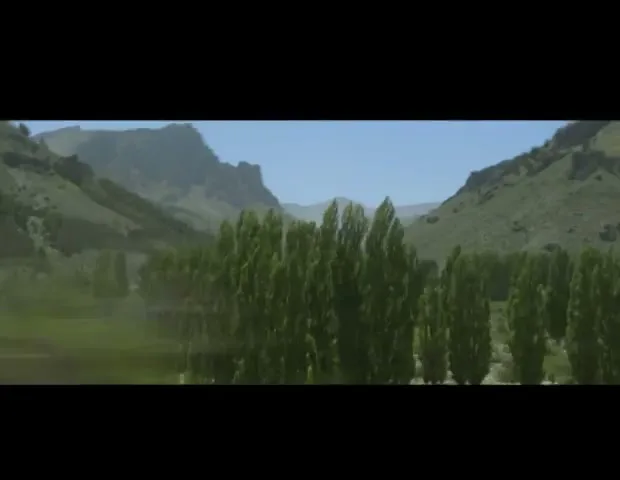 A wide shot of a green valley with trees and mountains under a clear blue sky, as if seen from a moving vehicle.