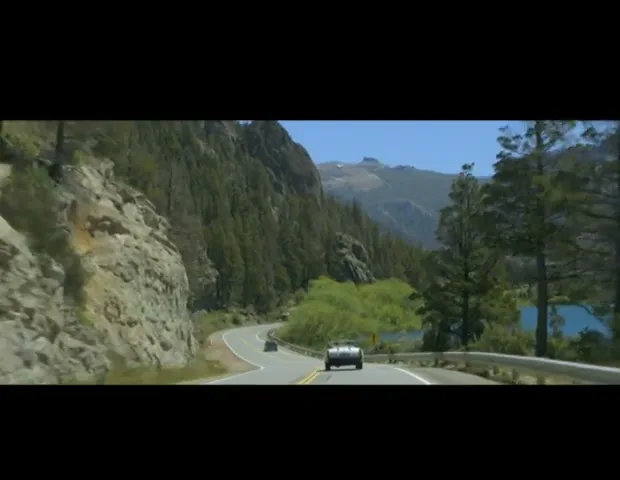 A silver car drives on a winding mountain road next to a lake, with green trees and rocky cliffs under a clear sky.