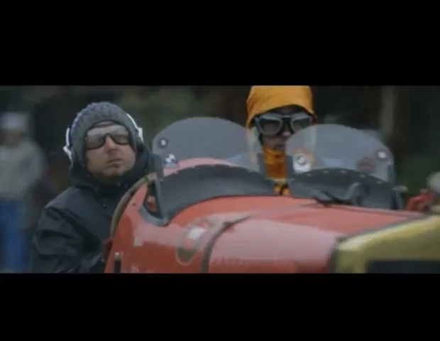 Two men in vintage racing gear push a red vintage race car on a wet road, with trees in the background.