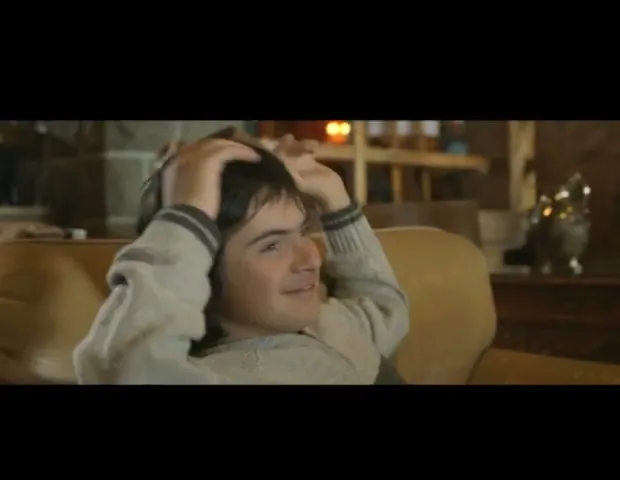 A young boy with dark hair, wearing a light-colored sweater, smiles and puts his hands behind his head while sitting on a couch in a cozy, rustic living room.