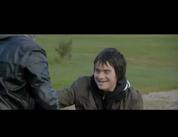 A young man with Down syndrome, wearing a dark jacket, laughs joyfully while holding onto another person's arm in a grassy outdoor setting.
