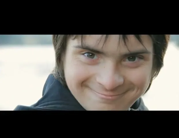 A young boy with dark hair and a blue jacket smiles mischievously at the camera outdoors.