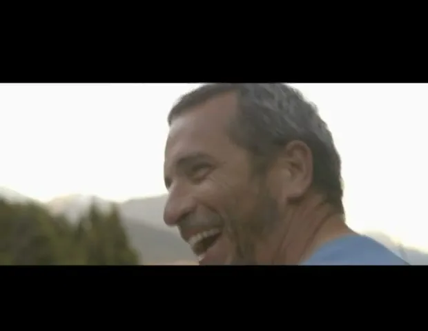 A man with graying hair and a beard laughs heartily, looking off-camera with mountains and trees in the background during the day.