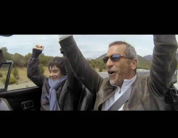 A man and a boy in a convertible car, driving on a road with mountains in the background, both raising their arms in excitement.